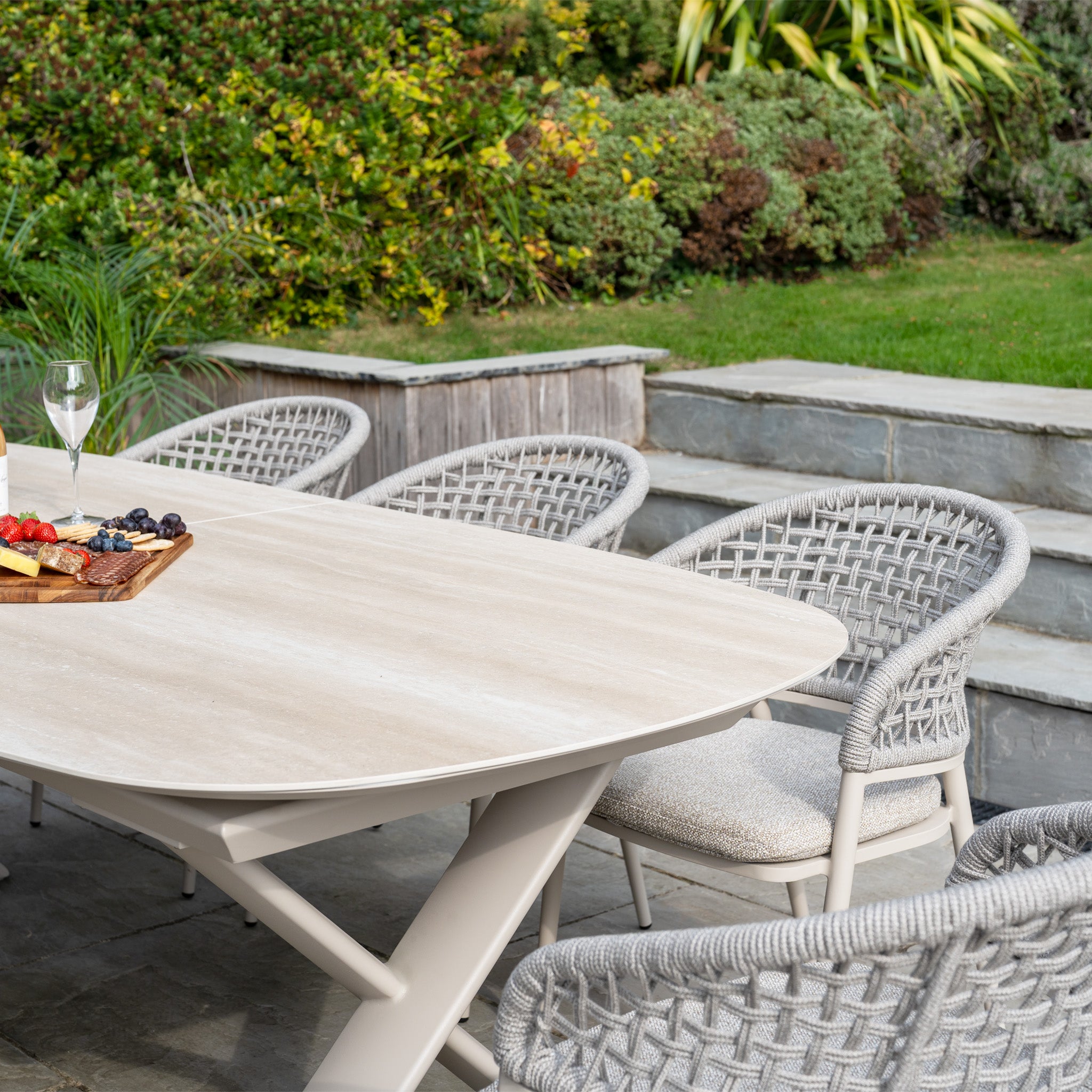 A patio table with a fruit and cheese platter, surrounded by woven chairs from the Kalama 8 Seat Oval Rope Dining Set with Albatross Extending Table in Oatmeal, creates an ideal outdoor dining spot in a lush garden.