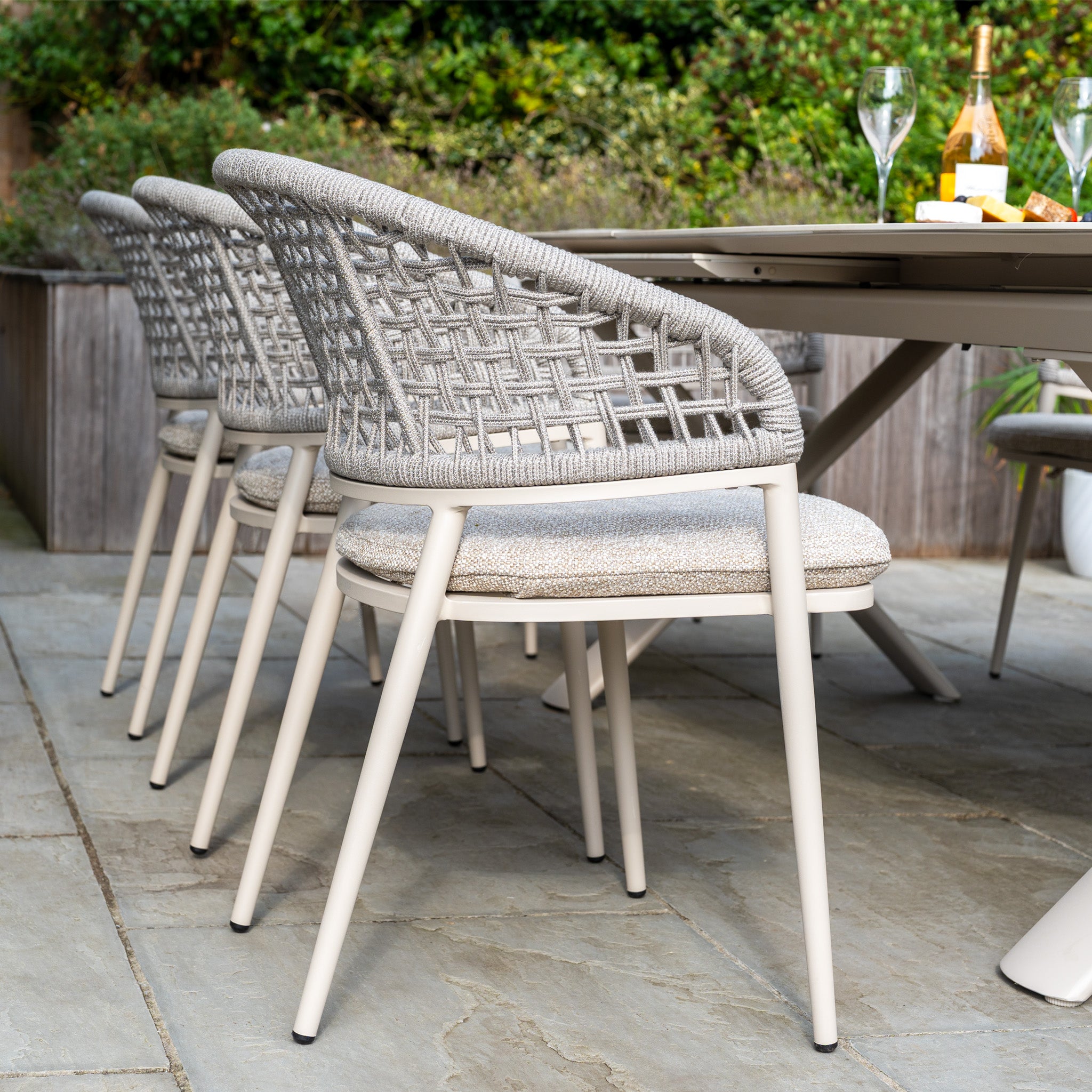 The Kalama 8 Seat Oval Rope Dining Set with Albatross Extending Table in Oatmeal, set with wine and glasses on a stone patio surrounded by greenery, is perfect for your next outdoor dining experience.