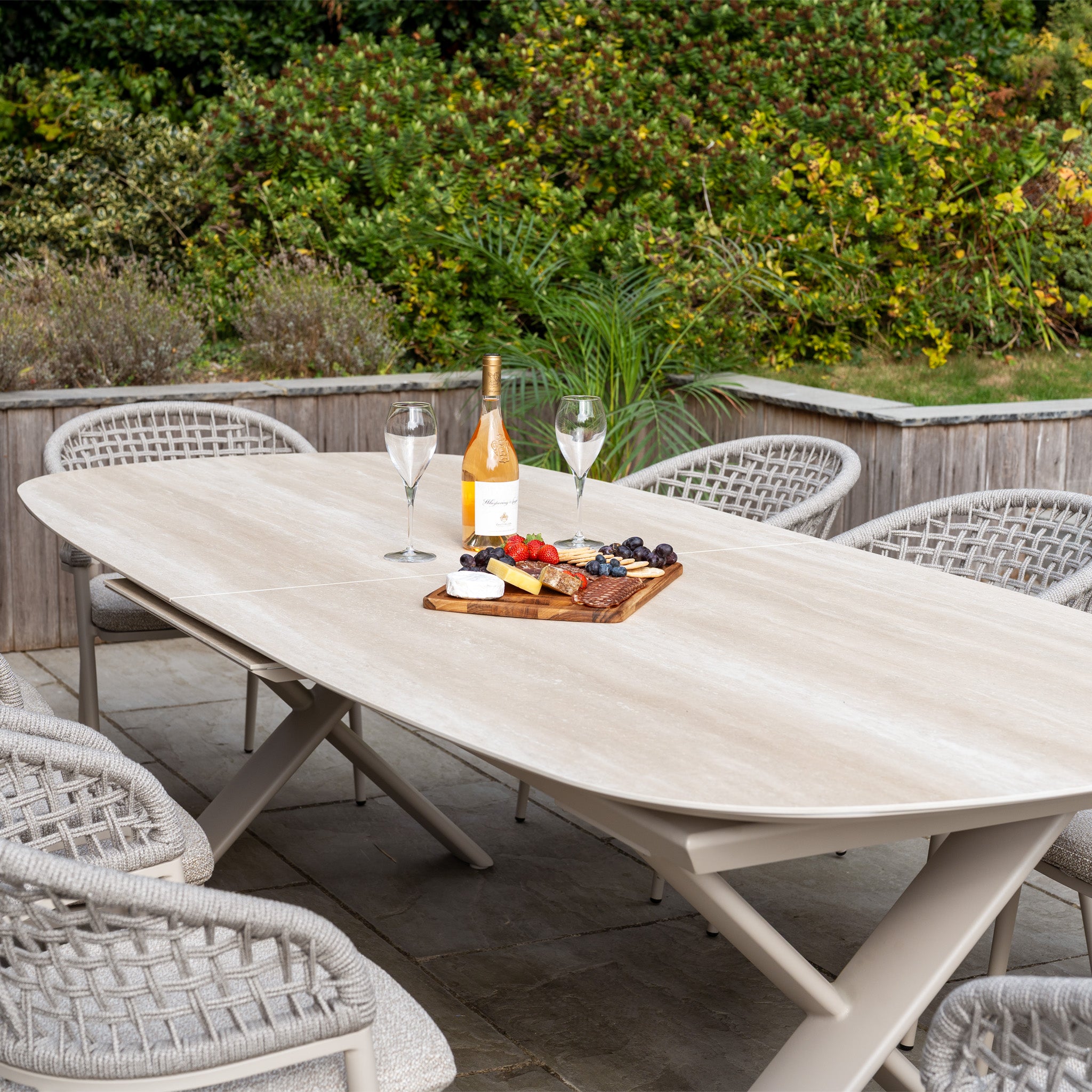 Enjoy elegant outdoor dining with the Kalama 8 Seat Oval Rope Dining Set, featuring the Albatross Extending Table in Oatmeal—perfect for gatherings with wine, glasses, charcuterie, and friends surrounded by greenery.