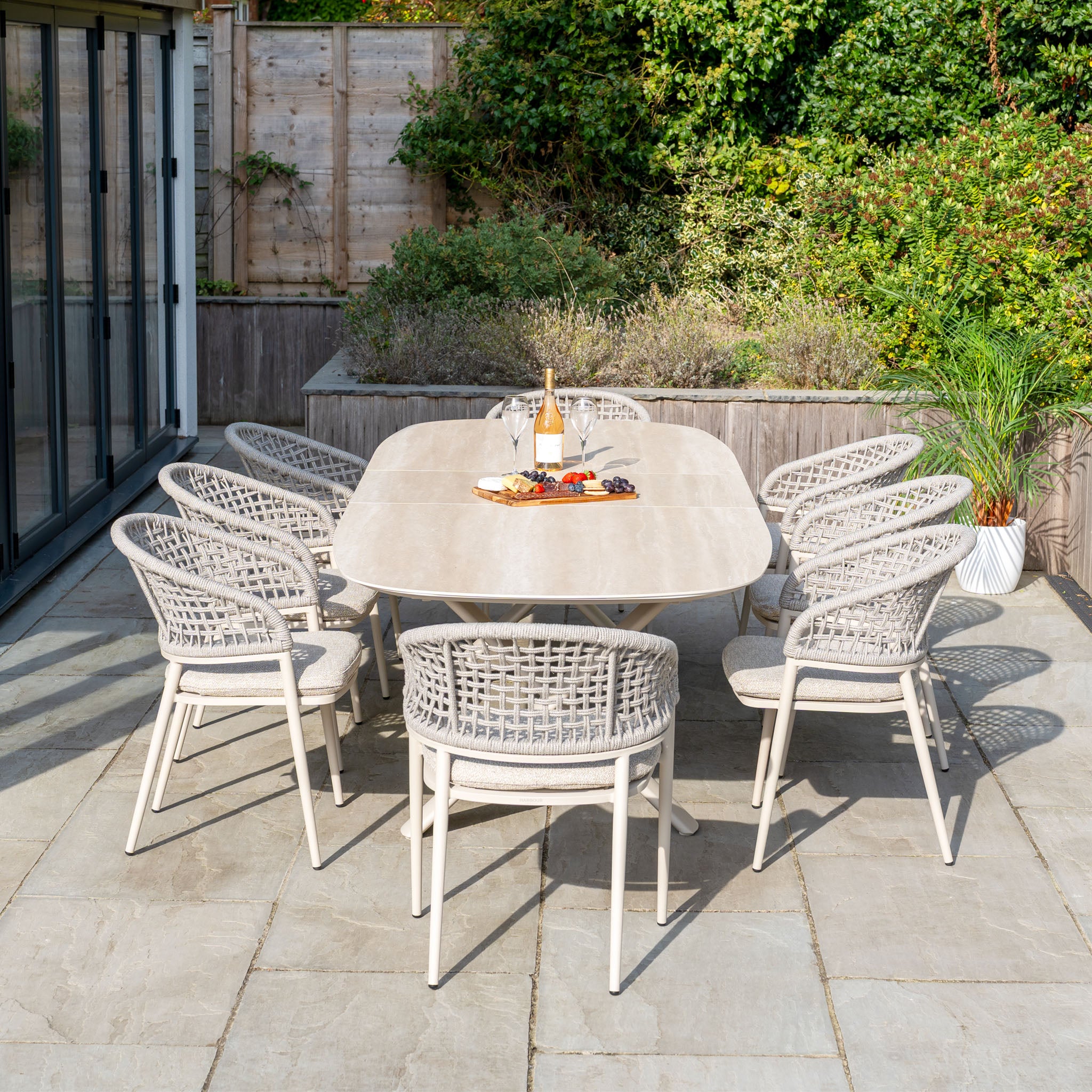 The Kalama 8 Seat Oval Rope Dining Set with Albatross Extending Table in Oatmeal is stylishly displayed with wine and snacks on a stone patio surrounded by a lush garden.