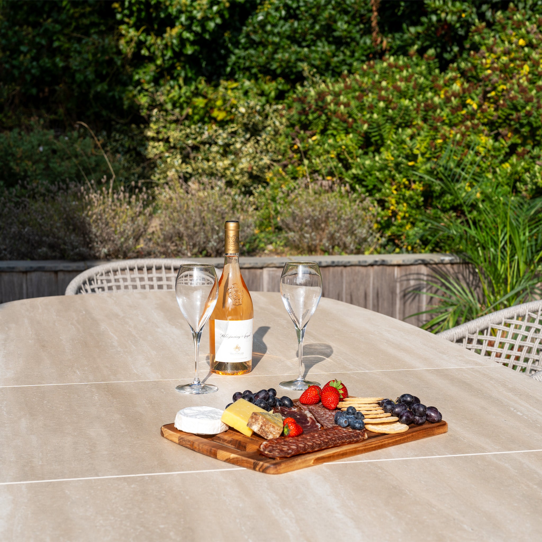 A bottle of wine, two glasses, and a cheese and fruit platter rest on the Kalama 8 Seat Oval Rope Dining Set with Albatross Extending Table in Oatmeal—perfect for relaxing evenings outdoors.