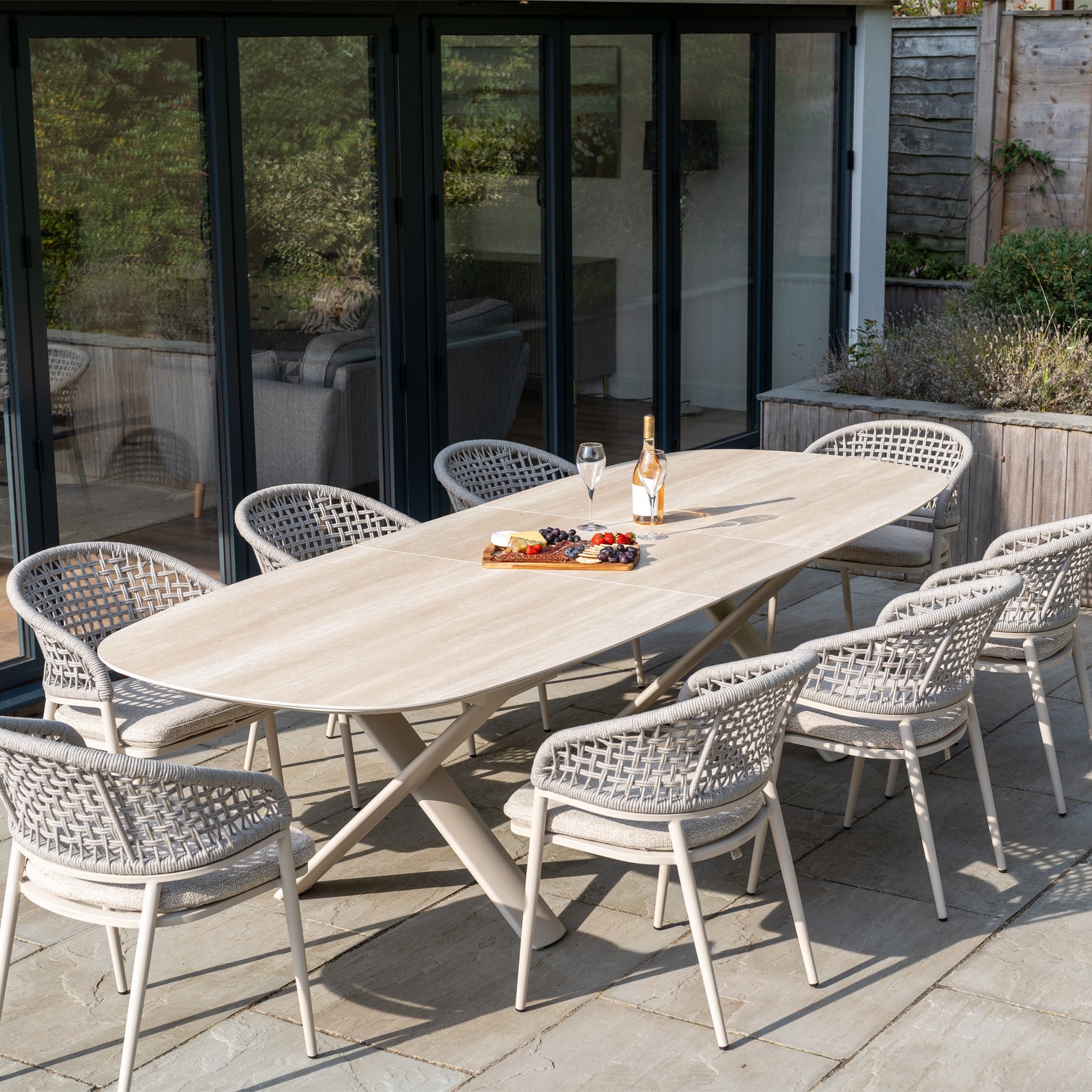 The Kalama 8 Seat Oval Rope Dining Set with Albatross Extending Table in Oatmeal is arranged on a stone patio near glass doors, complete with wine and snacks for outdoor dining.