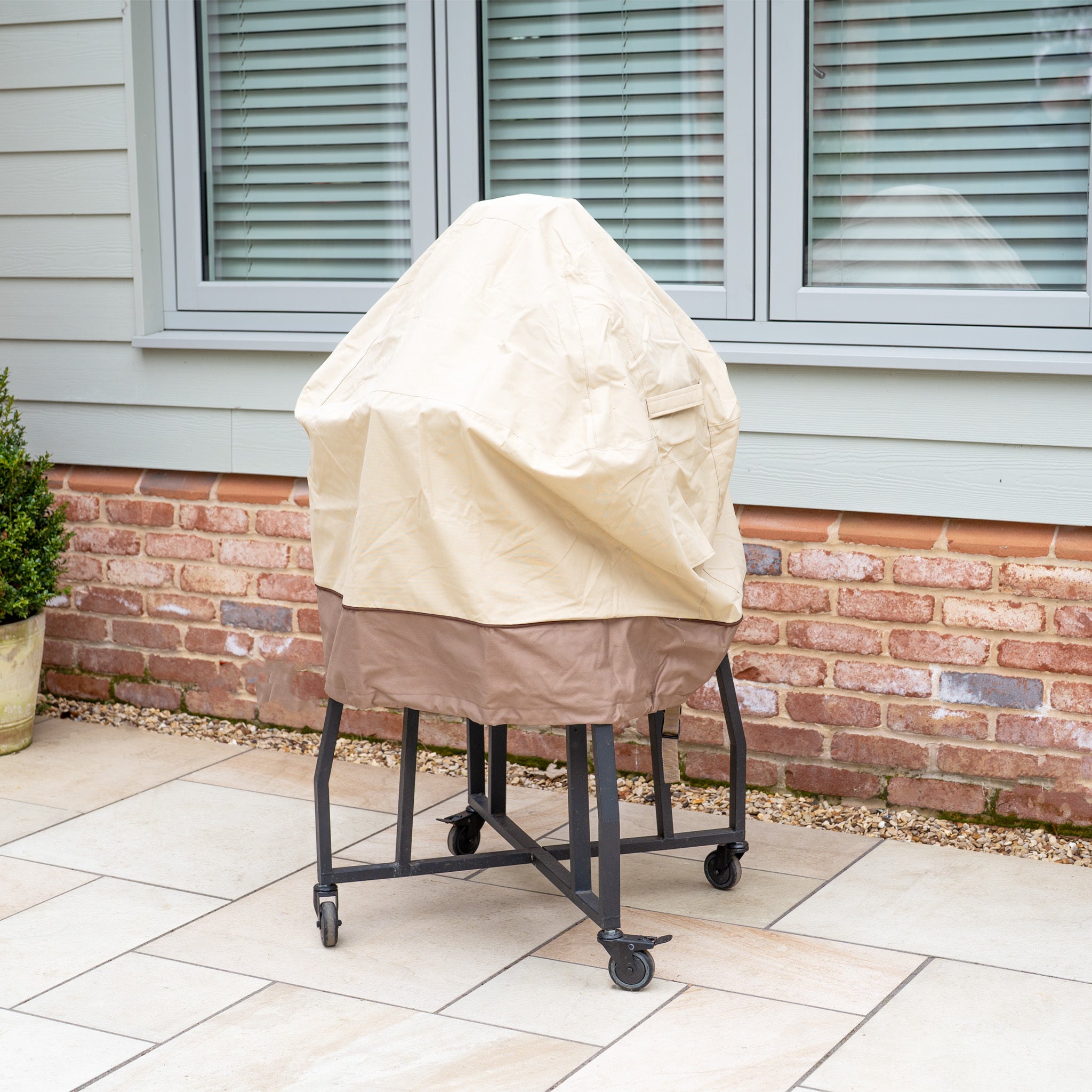 A covered outdoor grill sits on the patio, shielded by a Rain Cover for 22" Kamado Grills. The waterproof cover and wheels position it conveniently near a brick and siding wall, ensuring it’s protected against the elements.