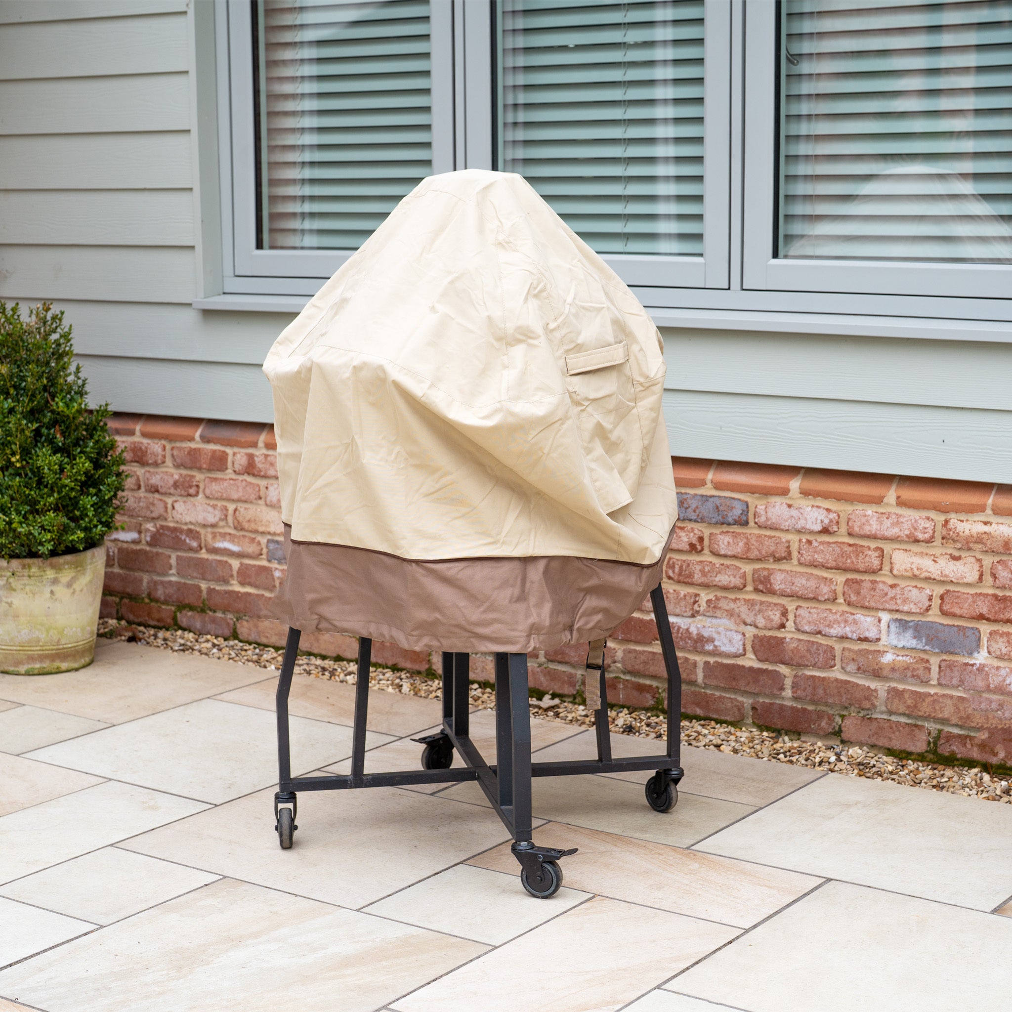 A 22" Kamado Grill is on the patio, covered with a beige Rain Cover for 22" Kamado Grills, near a healthy plant and window.