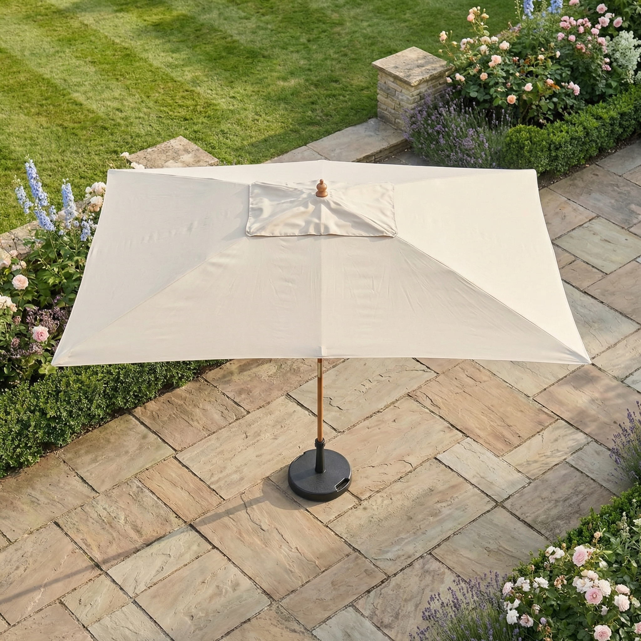 The Kratos 3m x 2m Rectangular Wooden Parasol in Beige (UV50+) stands on a stone terrace amid green grass and blooming garden flowers.
