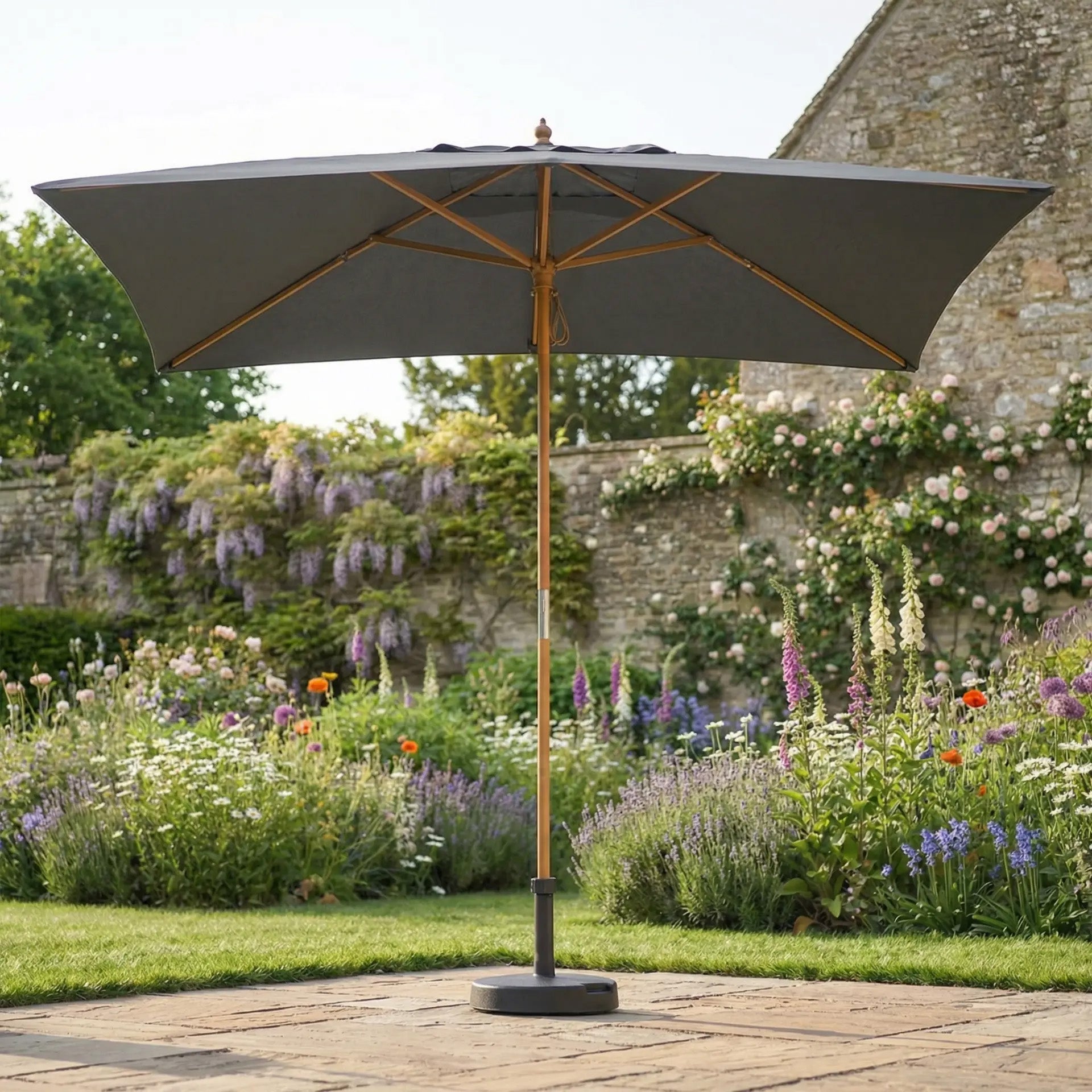 The Kratos 3m x 2m Rectangular Wooden Parasol in Grey stands on a stone patio, surrounded by blooming flowers and greenery, providing ideal sun protection for relaxing outdoors.