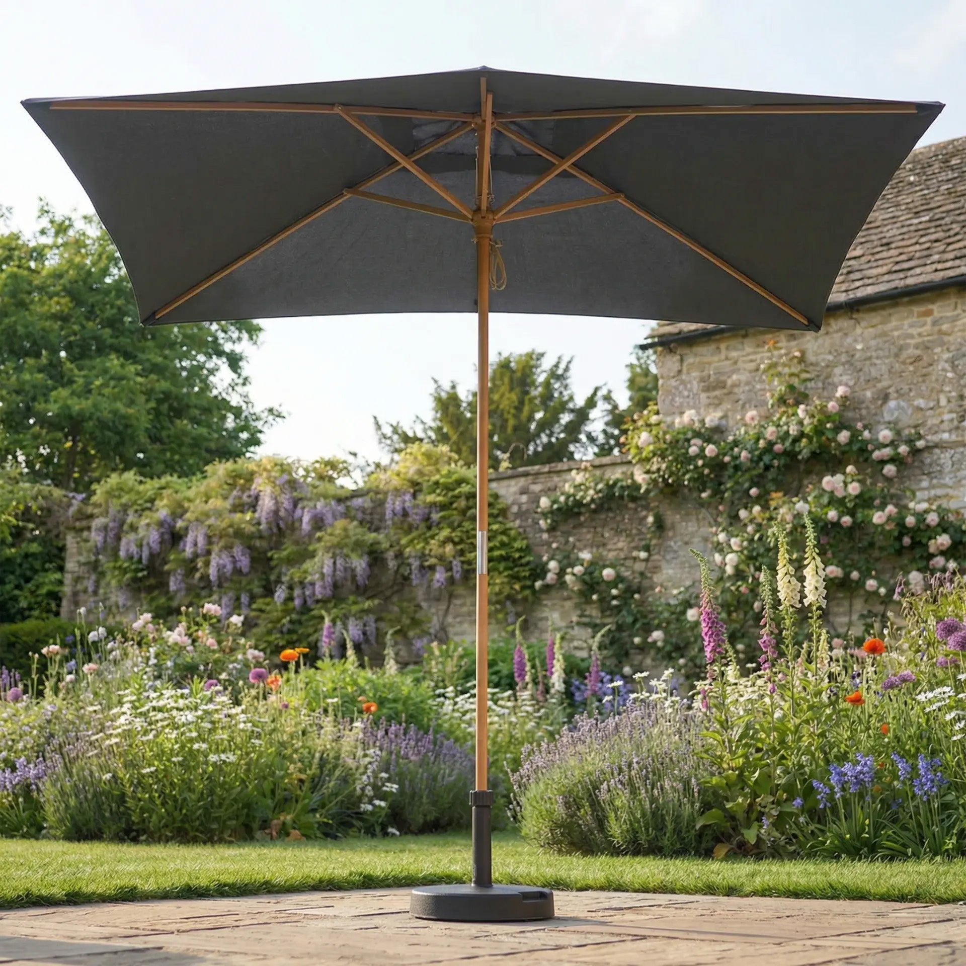 The Kratos 3m x 2m Rectangular Wooden Parasol in Grey stands on a stone terrace in a vibrant garden, providing elegant sun protection.