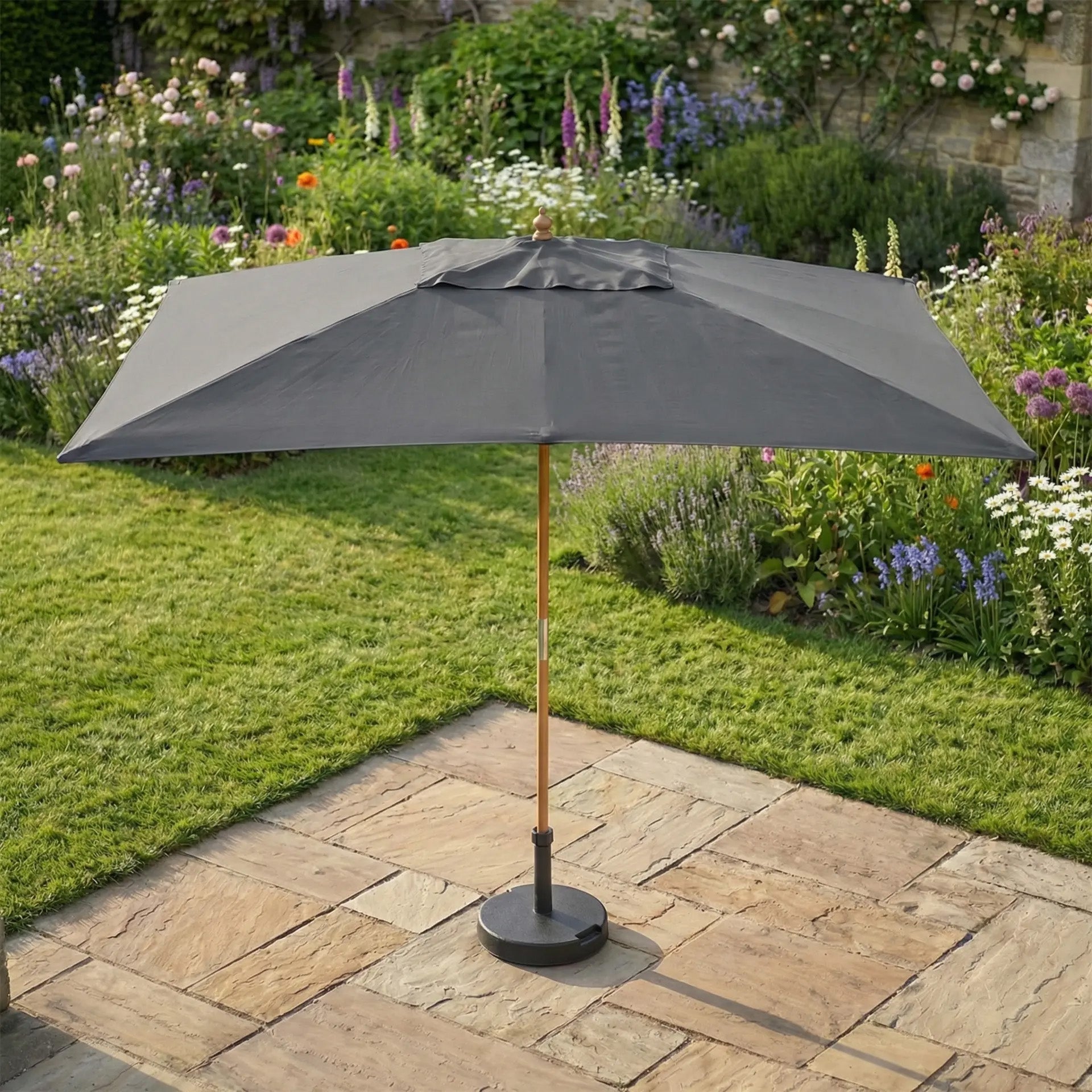 The Kratos 3m x 2m Rectangular Wooden Parasol in Grey stands on a stone patio, offering stylish sun protection in a lush garden brimming with vibrant flowers and greenery.