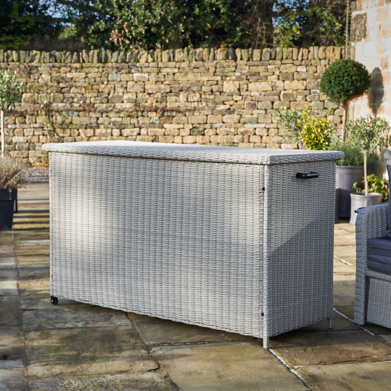 The Pacific Lifestyle Large Rattan Storage Box in Stone Grey sits perfectly on a stone patio, surrounded by a brick wall and lush greenery, ideal for outdoor cushion storage.