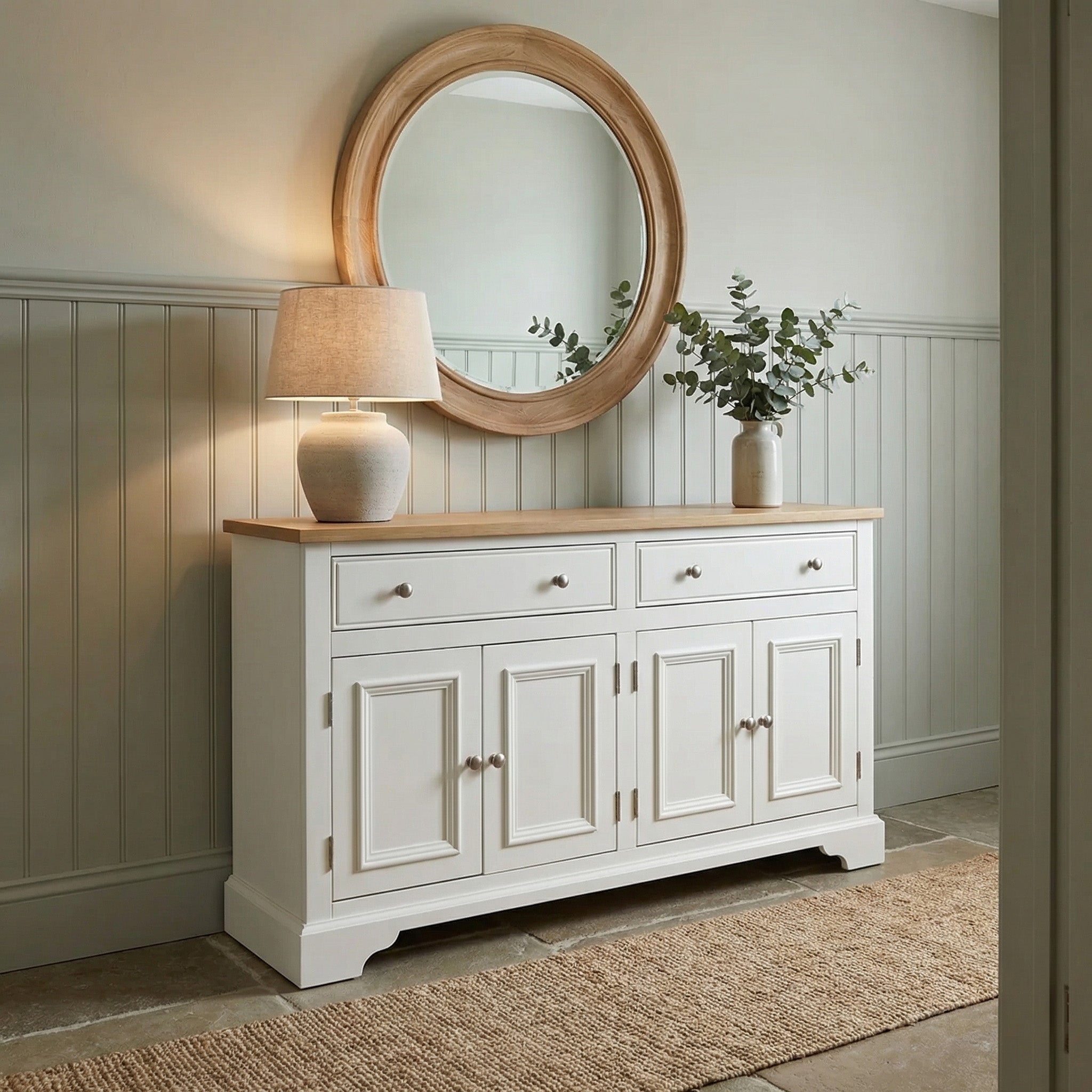 The Amberley Large 4 Door 2 Drawers Sideboard in Warm White stands in a softly lit, paneled hallway, topped with a lamp, green vase, and round mirror above.