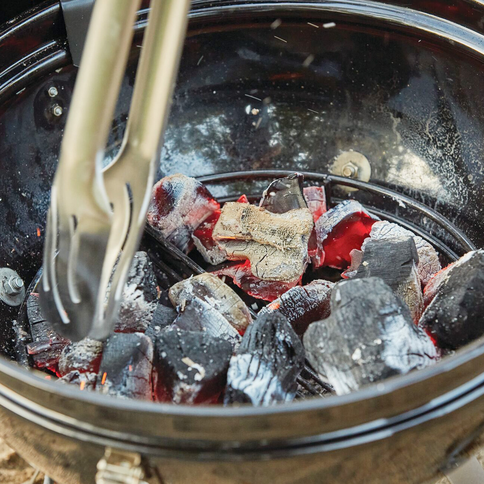 Close-up of tongs holding a charcoal piece in the Napoleon NK18 Charcoal Kettle Grill with glowing embers, highlighting the adjustable vents for precise control.