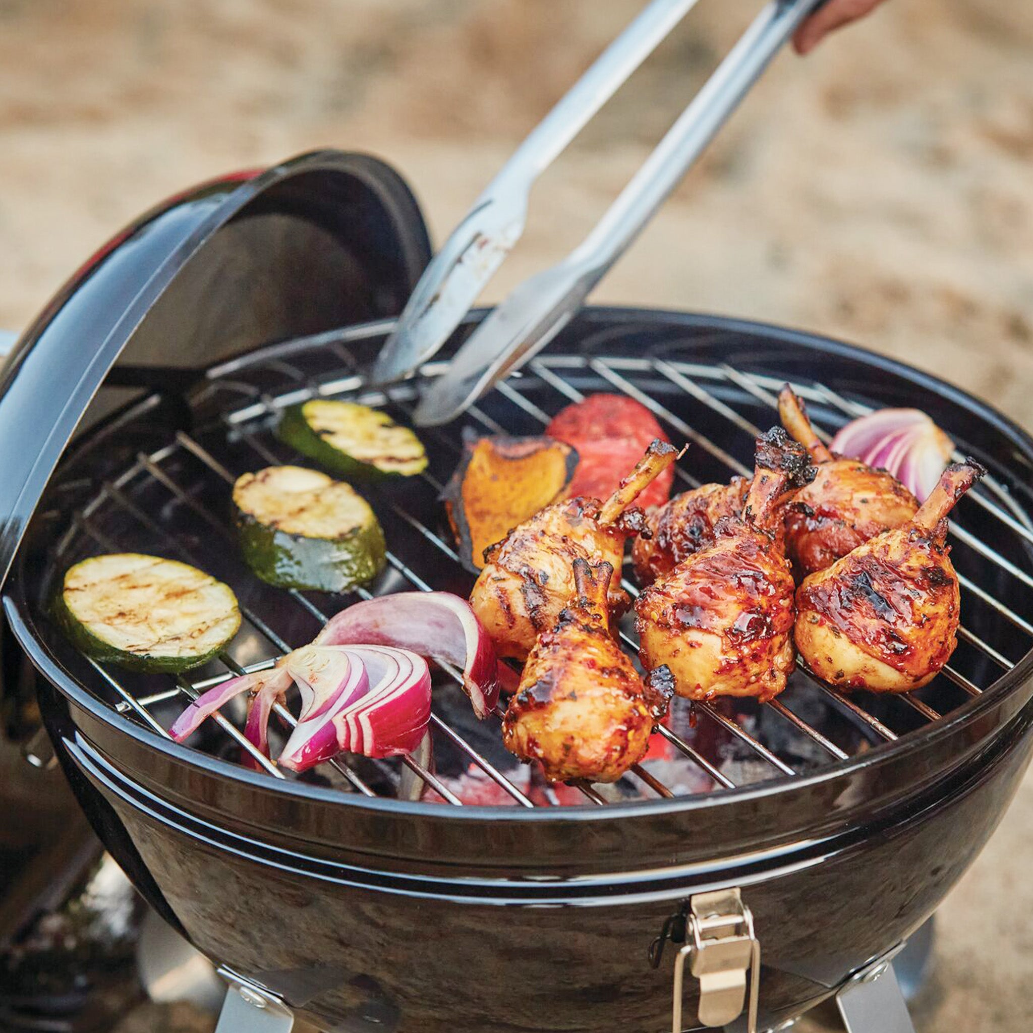 Grilled chicken drumsticks and vegetables are being cooked on the Napoleon NK18 Charcoal Kettle Grill, with a pair of tongs adjusting the food. The adjustable vents and ACCU-PROBE™ Temperature Gauge ensure perfect cooking control.