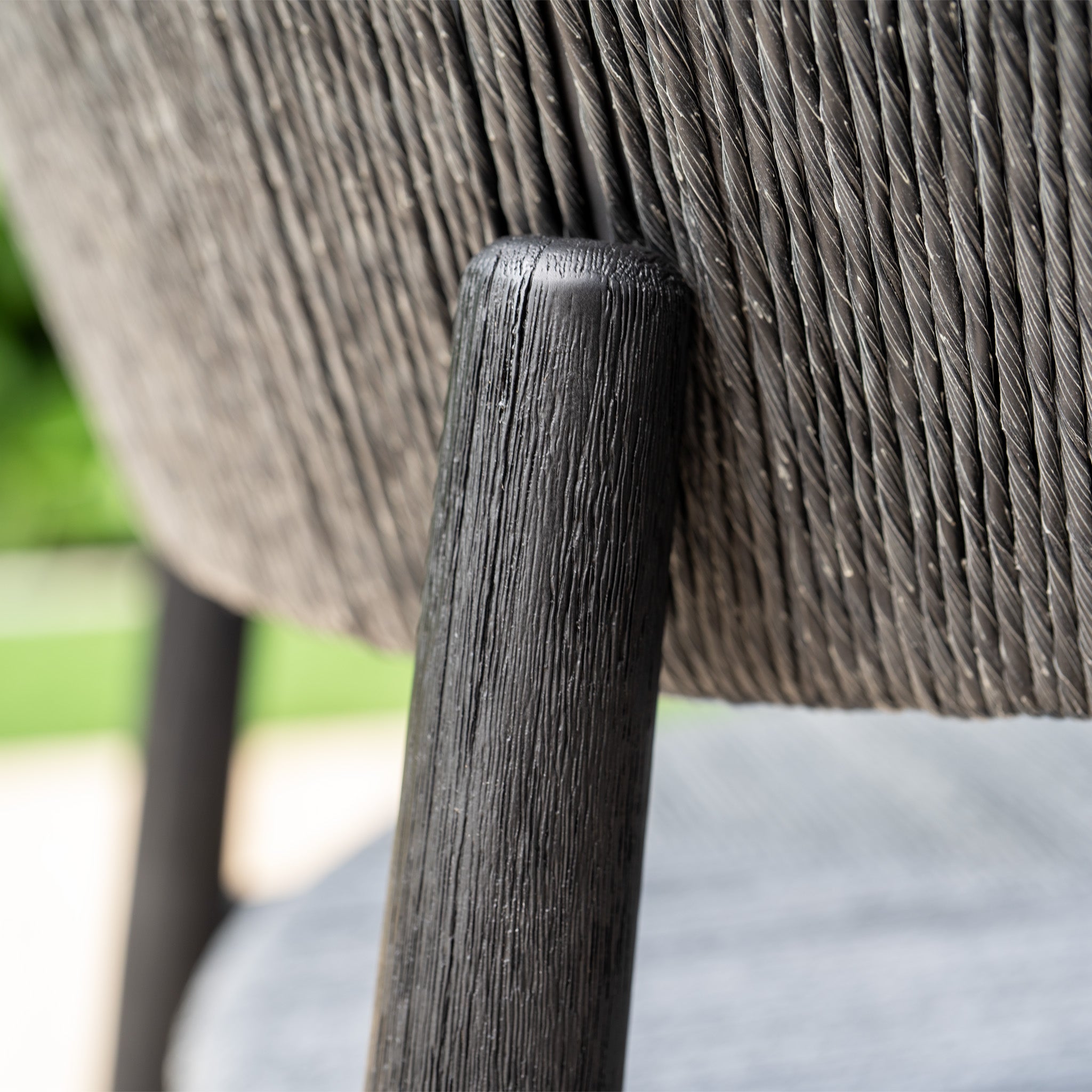 Close-up of a textured, dark wooden chair leg and hand-woven wicker backrest, shown outdoors—ideal for pairing with the Lucca 6 Seat Dining Set with Teak Round Table in Black.