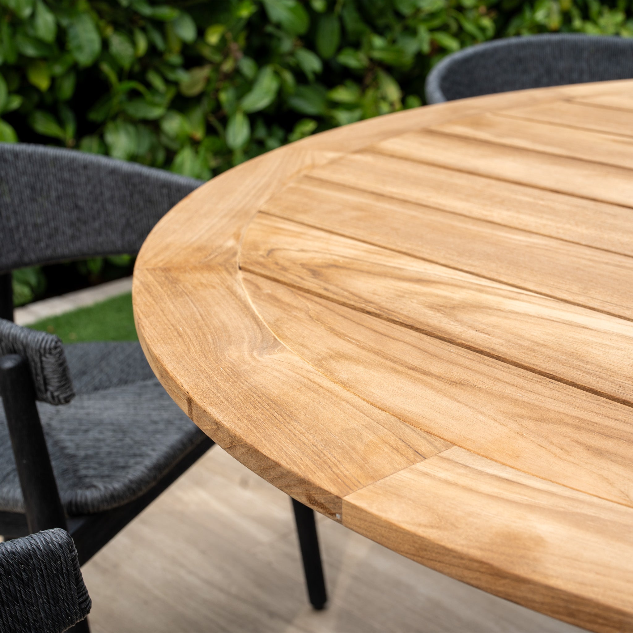 The Lucca 6 Seat Dining Set features a stylish teak round table in black, ideal for outdoor dining amid green foliage, offering an inviting and comfortable setting.