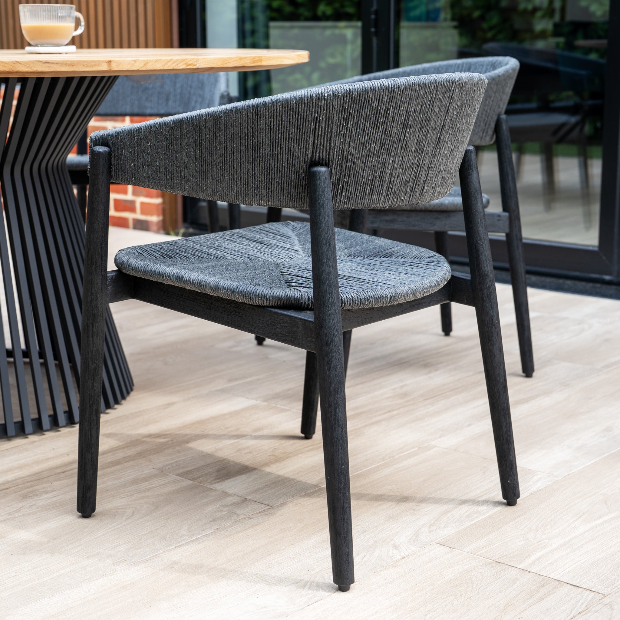The Lucca 6 Seat Dining Set features black hand-woven wicker chairs paired with a round teak table, perfect for modern dining spaces.