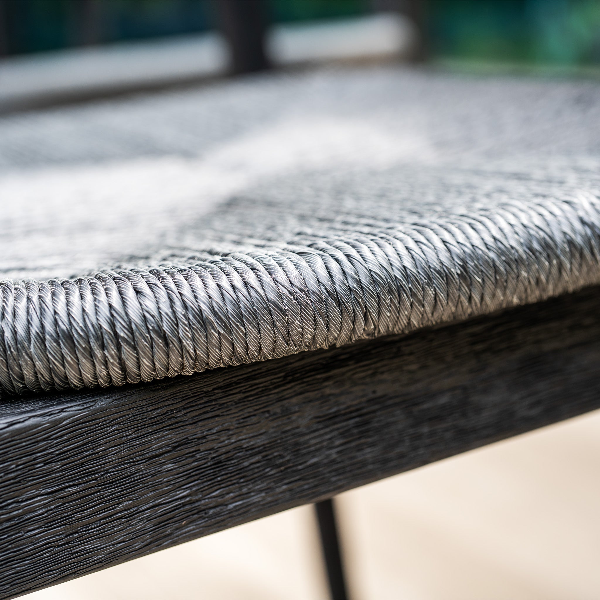Close-up of the Lucca 6 Seat Dining Set's hand-woven gray rattan chair seat with dark wood frame, shown in natural light alongside its teak round table in black.
