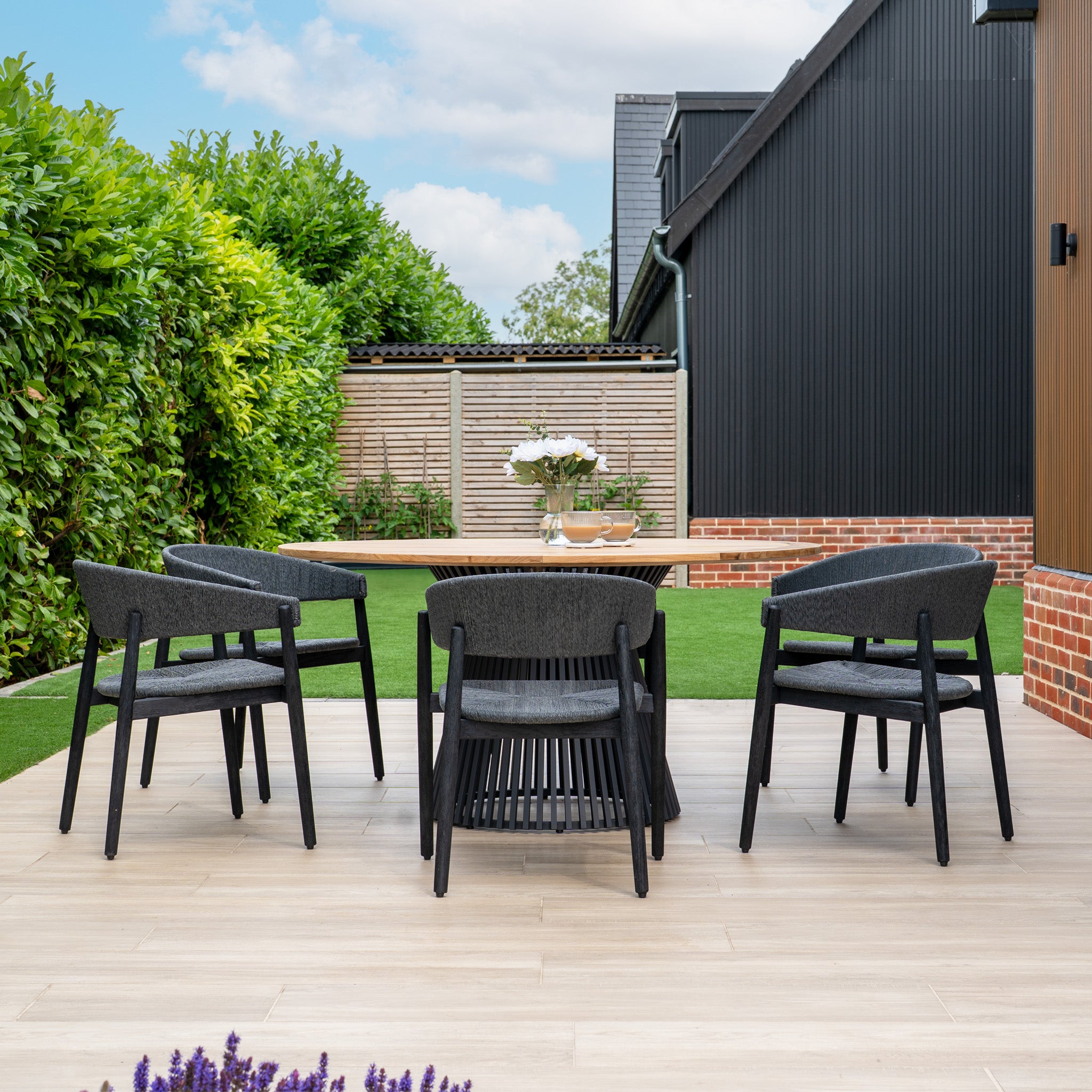 The Lucca 6 Seat Dining Set features a teak round table and black chairs, creating an inviting outdoor dining area with lush greenery and a sleek black building in the background.