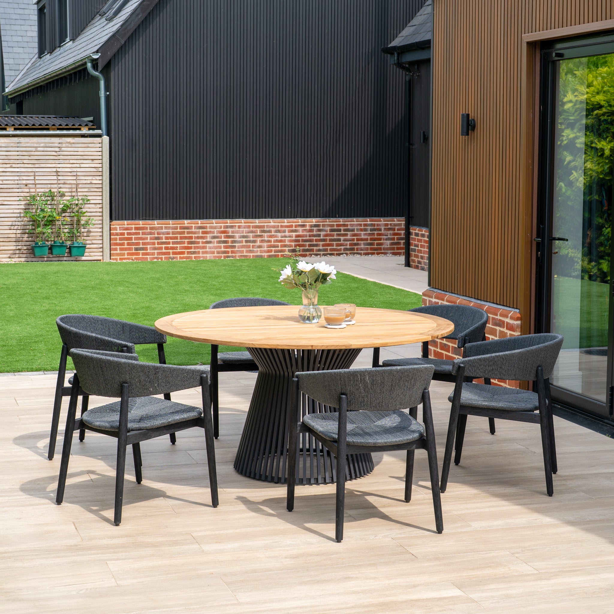 The Lucca 6 Seat Dining Set features a teak round table and six black chairs, offering a stylish and inviting dining area perfect for modern outdoor spaces.