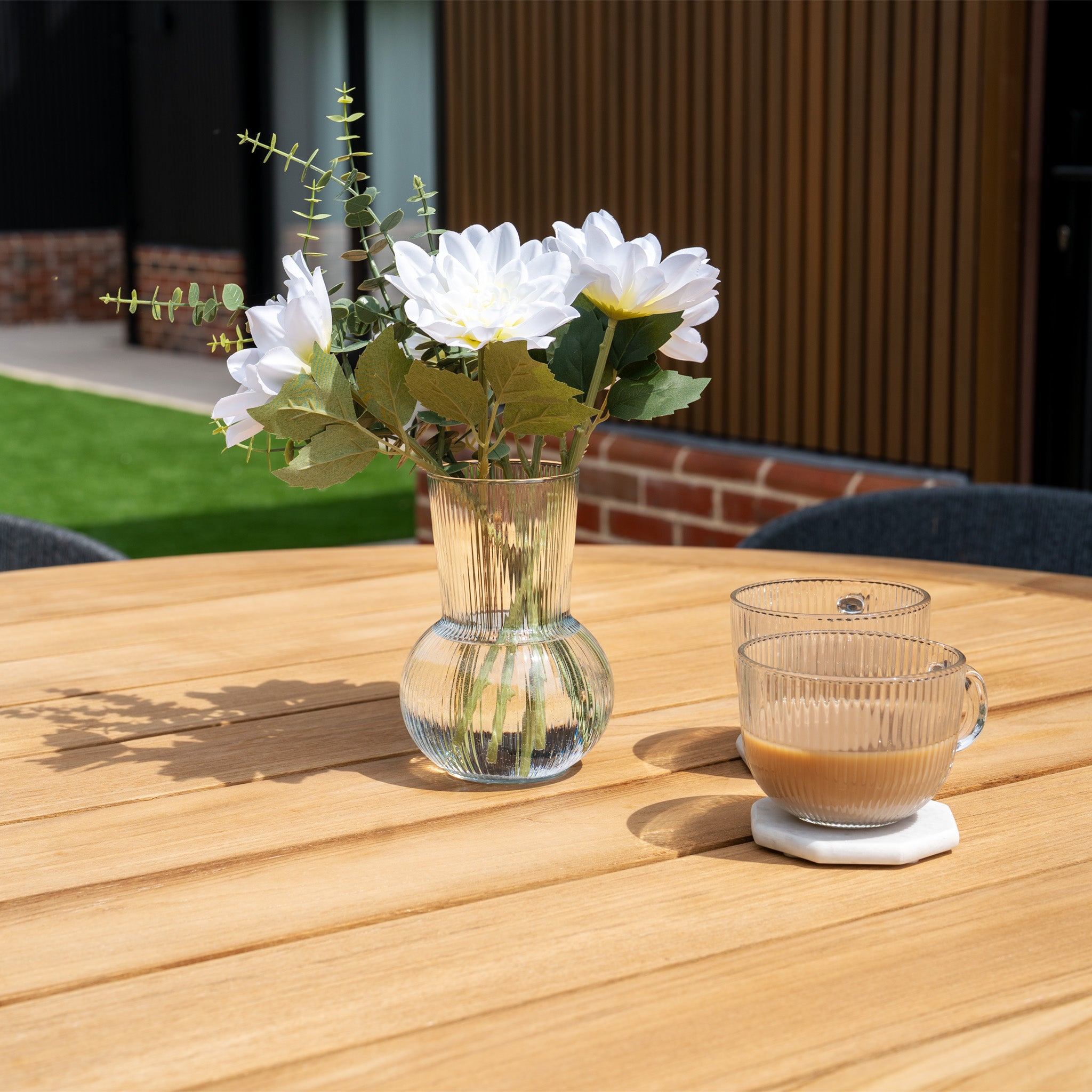 A glass vase with white flowers and two cups of coffee sit on the Lucca 6 Seat Dining Set with Teak Round Table in Black, glowing in the sunlight.