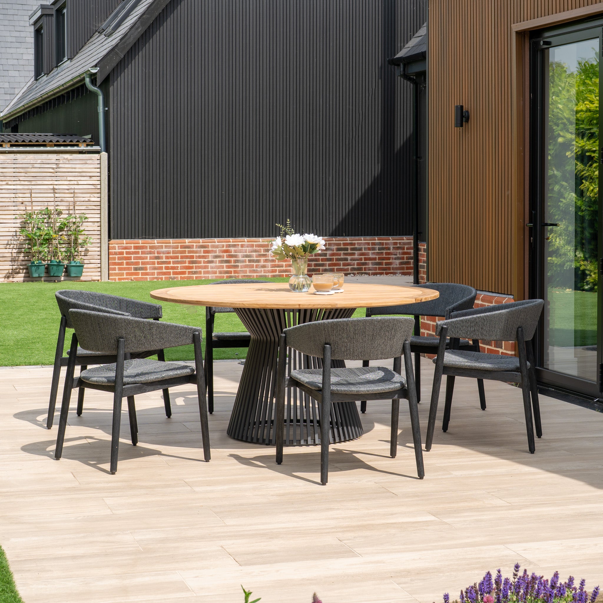 The Lucca 6 Seat Dining Set with Teak Round Table in Black, paired with four chairs and a floral vase, creates a stylish modern terrace setting perfect for outdoor living.