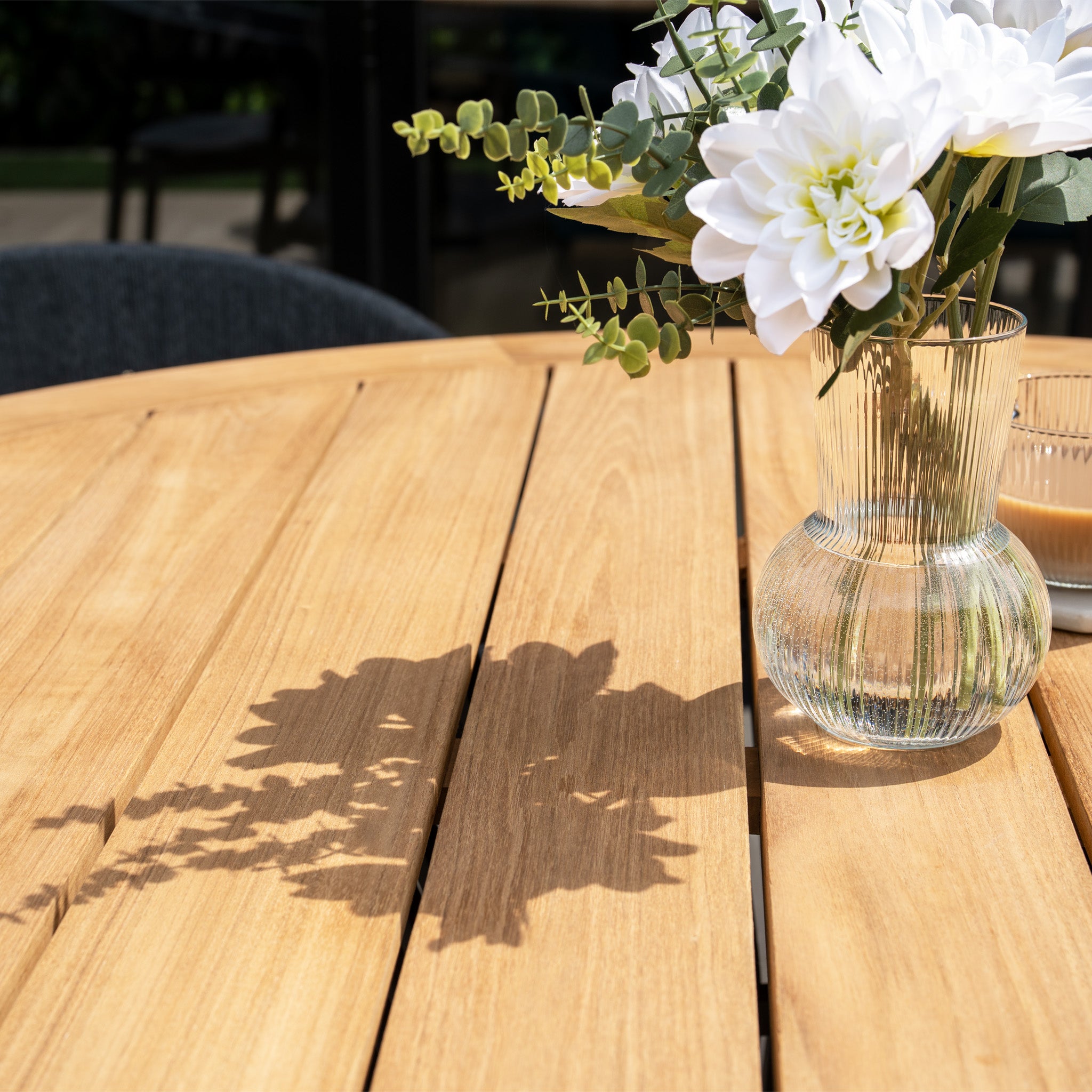 White flowers in a glass vase cast a shadow on the Lucca 6 Seat Dining Set with Teak Round Table in Black under sunlight.