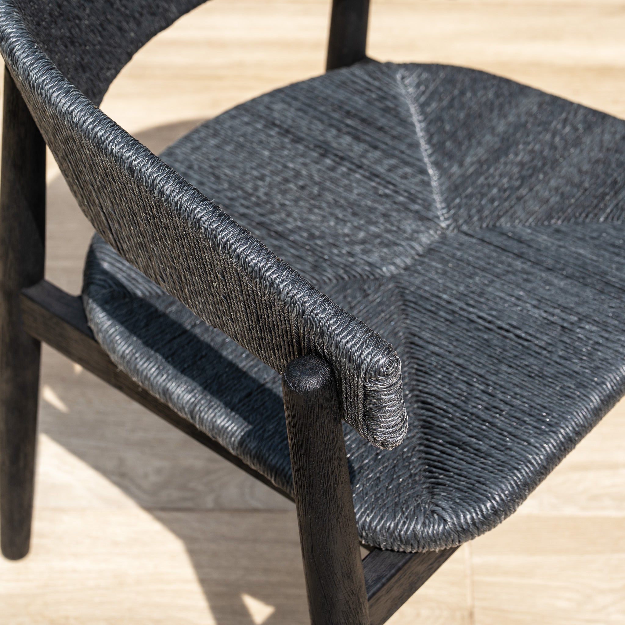 A close-up of the Lucca 6 Seat Dining Set with Teak Round Table in Black, featuring hand-woven geometric chairs on a light wooden floor—ideal for outdoor dining.