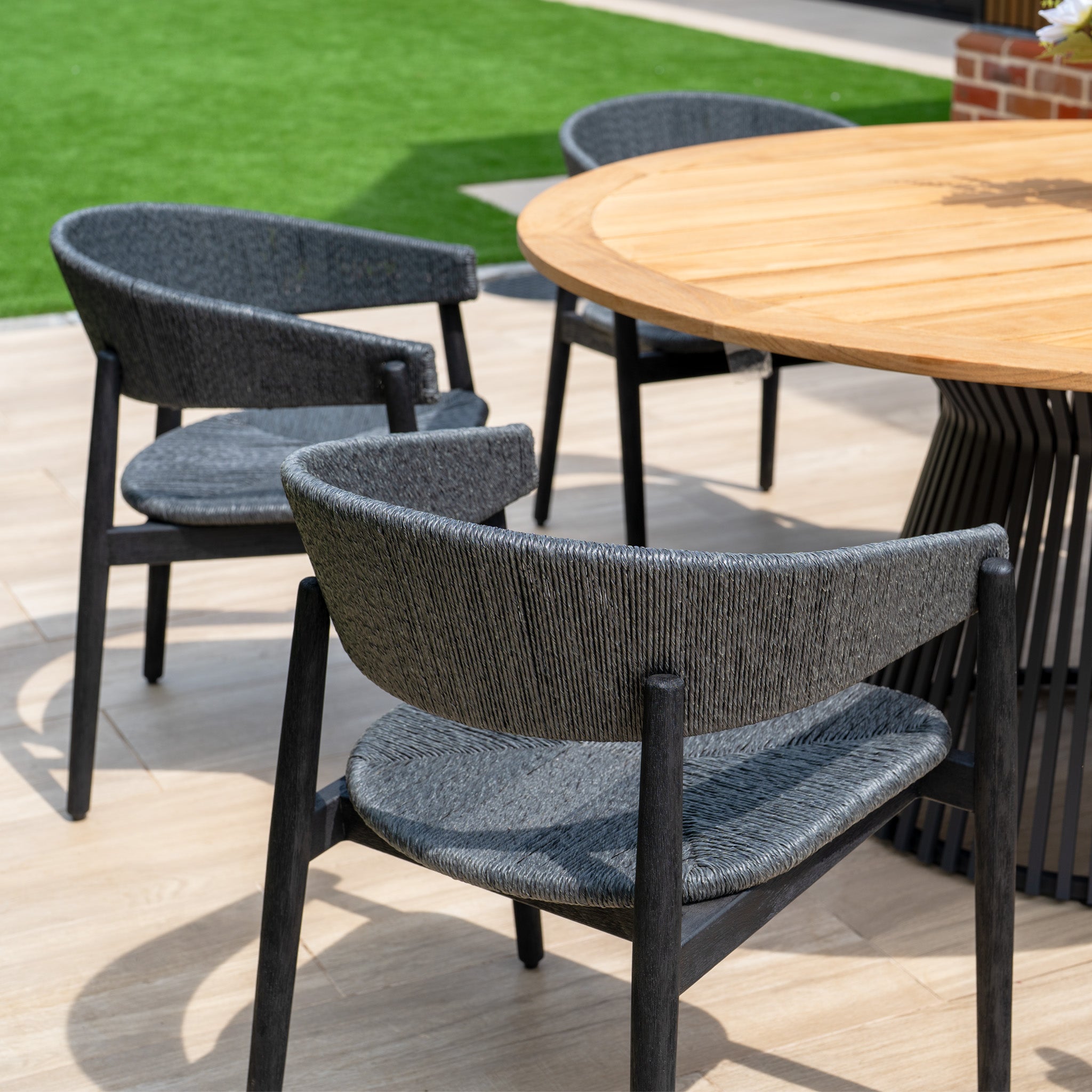 The Lucca 6 Seat Dining Set features a teak round table and black chairs, creating a stylish outdoor dining area on your patio, perfect for relaxing near artificial grass.