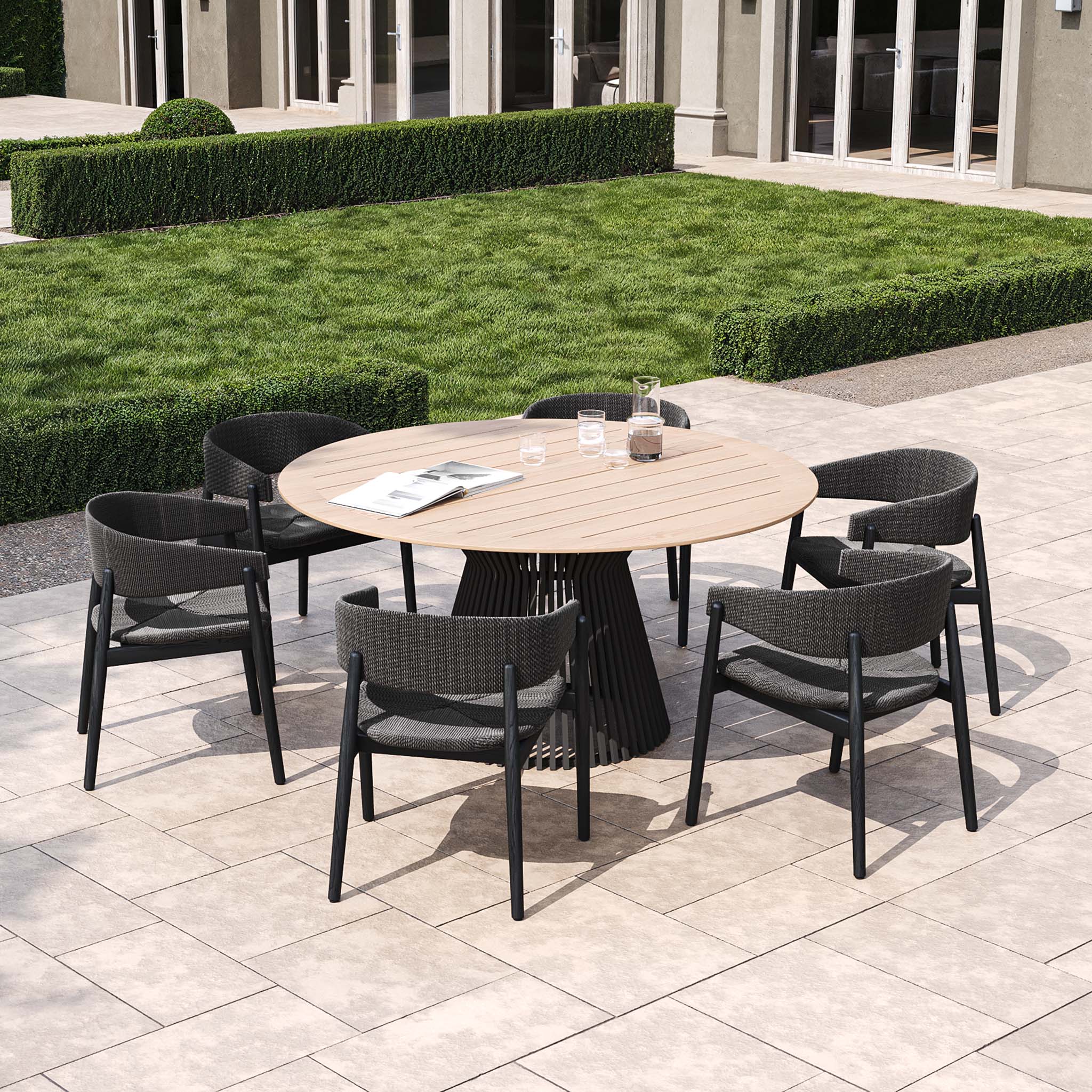 The Lucca 6 Seat Dining Set with a teak round table in black is ideal for elevating your outdoor living space, featuring on a tiled patio next to a manicured lawn and building.