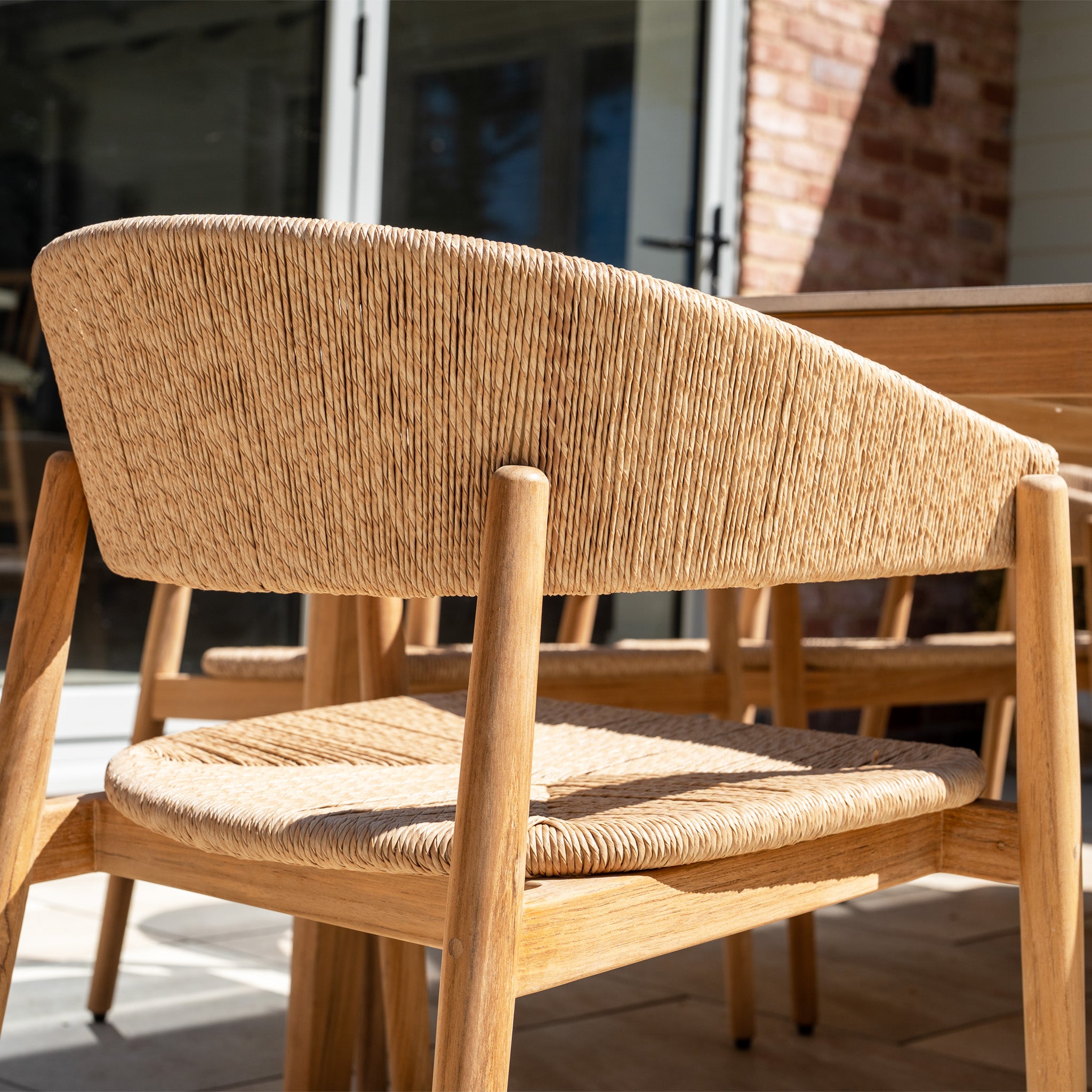 Close-up of a woven wooden chair from the Lucca 8 Seat Dining Set with Ceramic Top Table in Natural Weave, at an outdoor table with sunlight casting shadows on the patio.