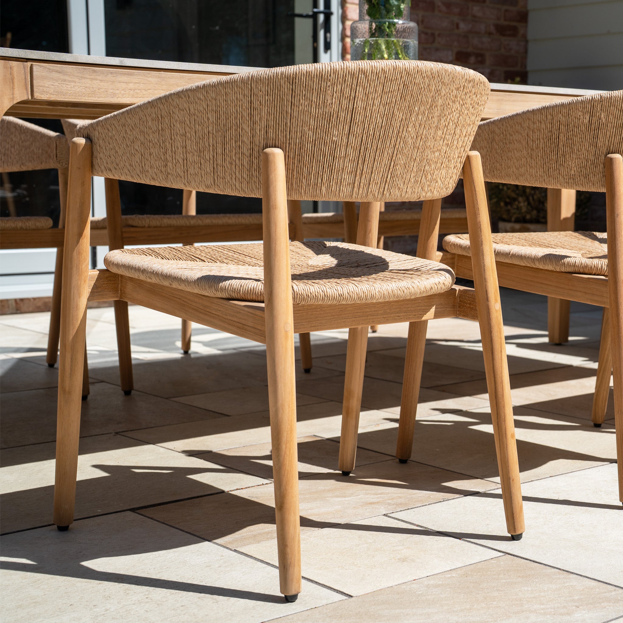A wooden chair with woven seat and curved backrest sits on a tiled patio, sunlight casting shadows—part of the Lucca 8 Seat Dining Set with Ceramic Top Table in Natural Weave, perfect for stylish outdoor settings.