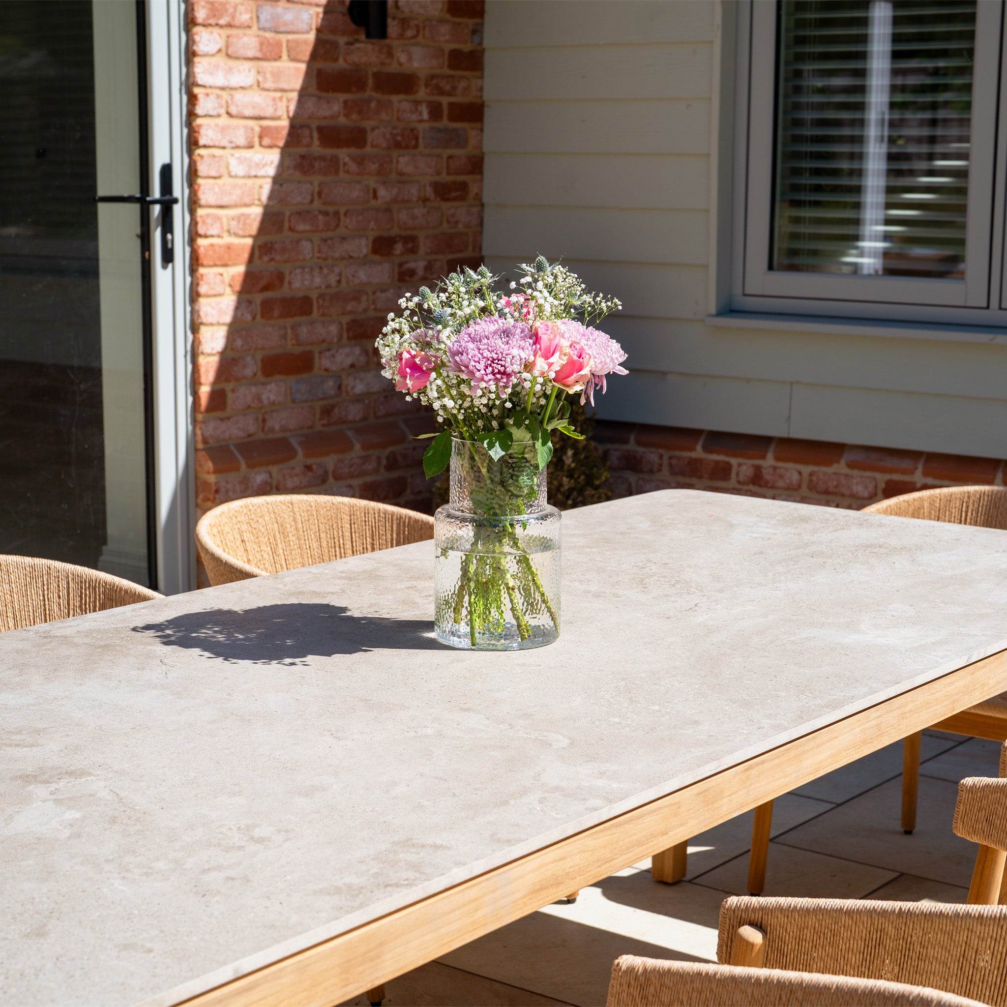 A vase of pink and white flowers sits on the sunlit Lucca 8 Seat Dining Set with Ceramic Top Table in Natural Weave, surrounded by wicker chairs—ideal outdoor furniture for gatherings.