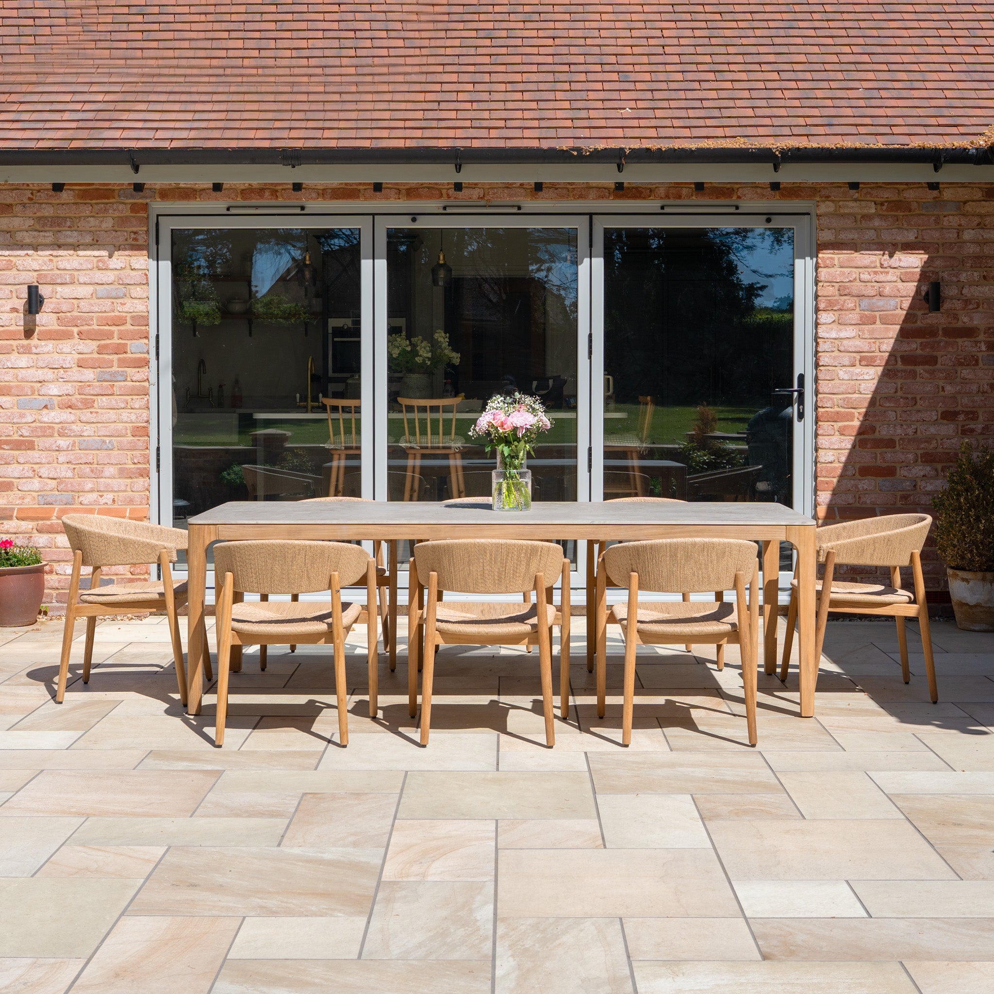 The Lucca 8 Seat Dining Set with Ceramic Top Table in Natural Weave offers a stylish wooden outdoor arrangement for eight, perfect for patios and enhancing spaces by a brick house.