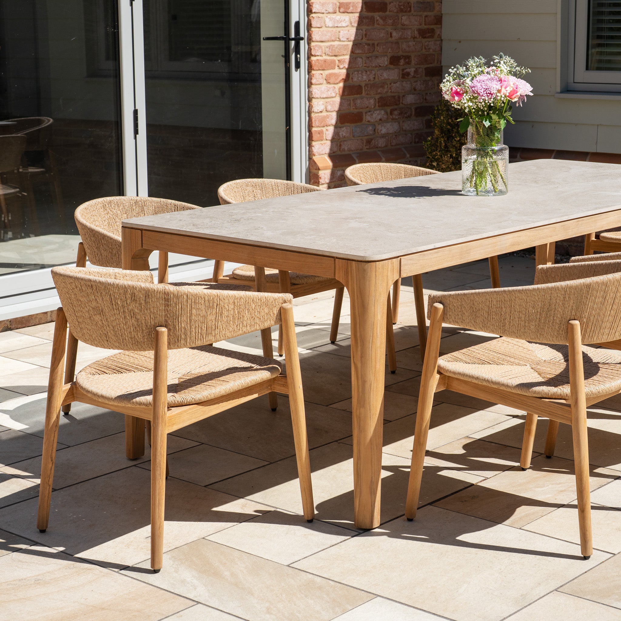 The Lucca 8 Seat Dining Set with Ceramic Top Table in Natural Weave features a stylish patio table and eight wicker chairs, arranged on a sunlit tiled terrace with a vase of flowers as the finishing touch.
