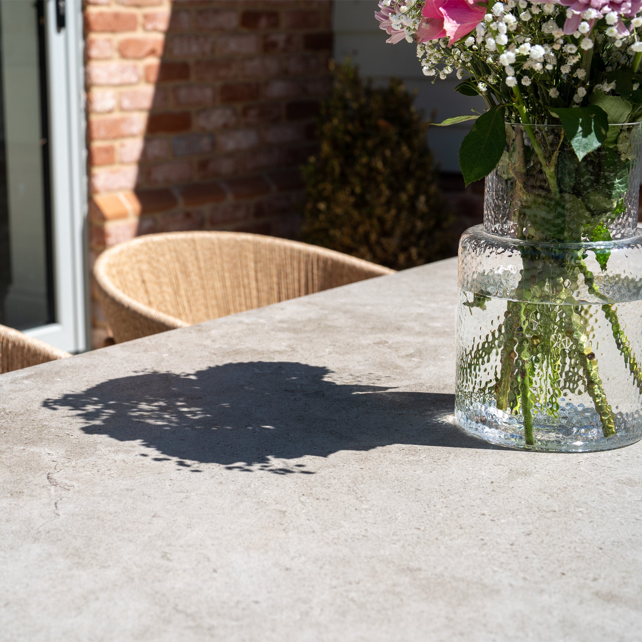 A vase of flowers casts a shadow on the sunlit Lucca 8 Seat Dining Set with Ceramic Top Table in Natural Weave, adding charm to your outdoor furniture arrangement.