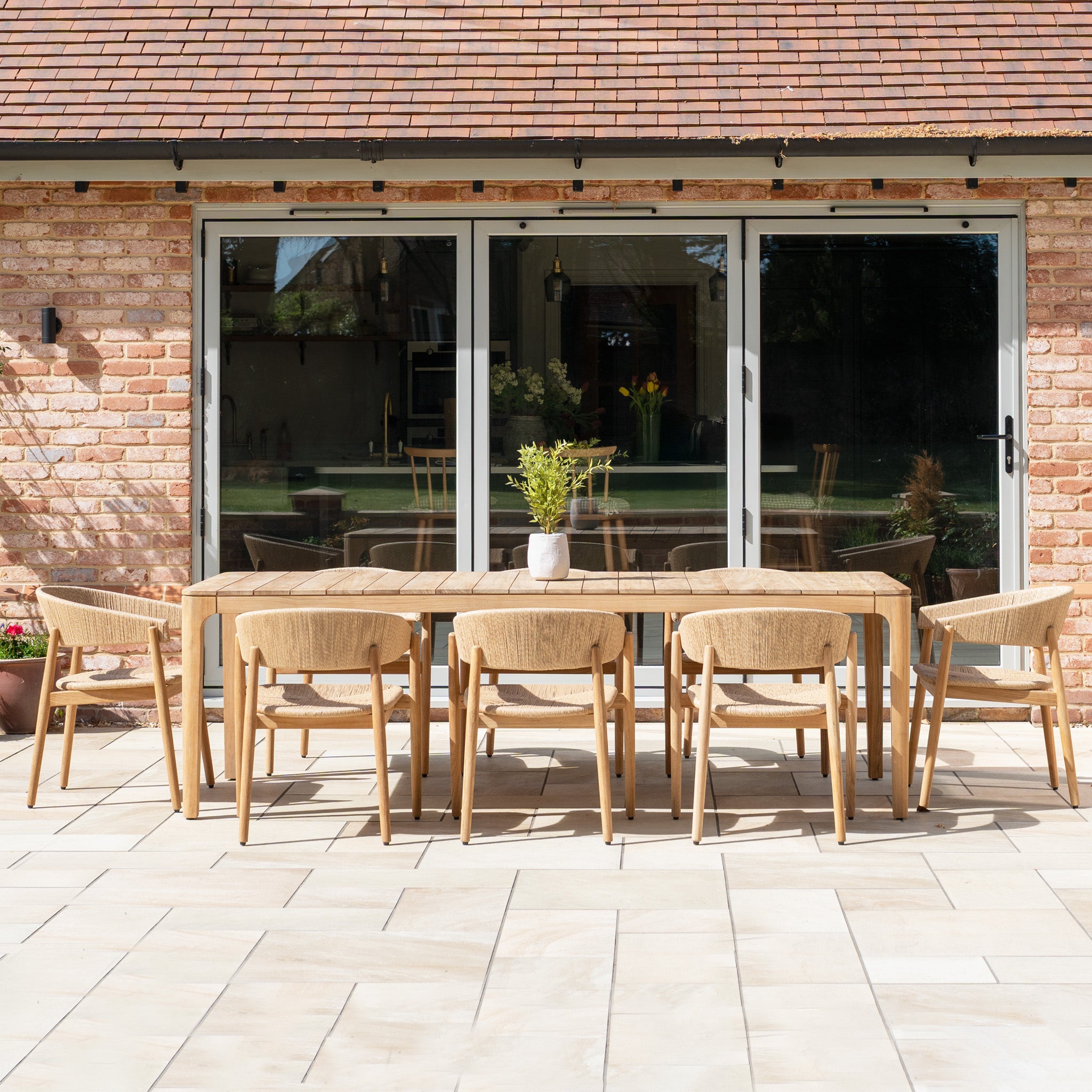 The Lucca 8 Seat Dining Set with Teak Table in Natural Weave creates an inviting patio dining area, perfect for outdoor meals in front of glass doors.