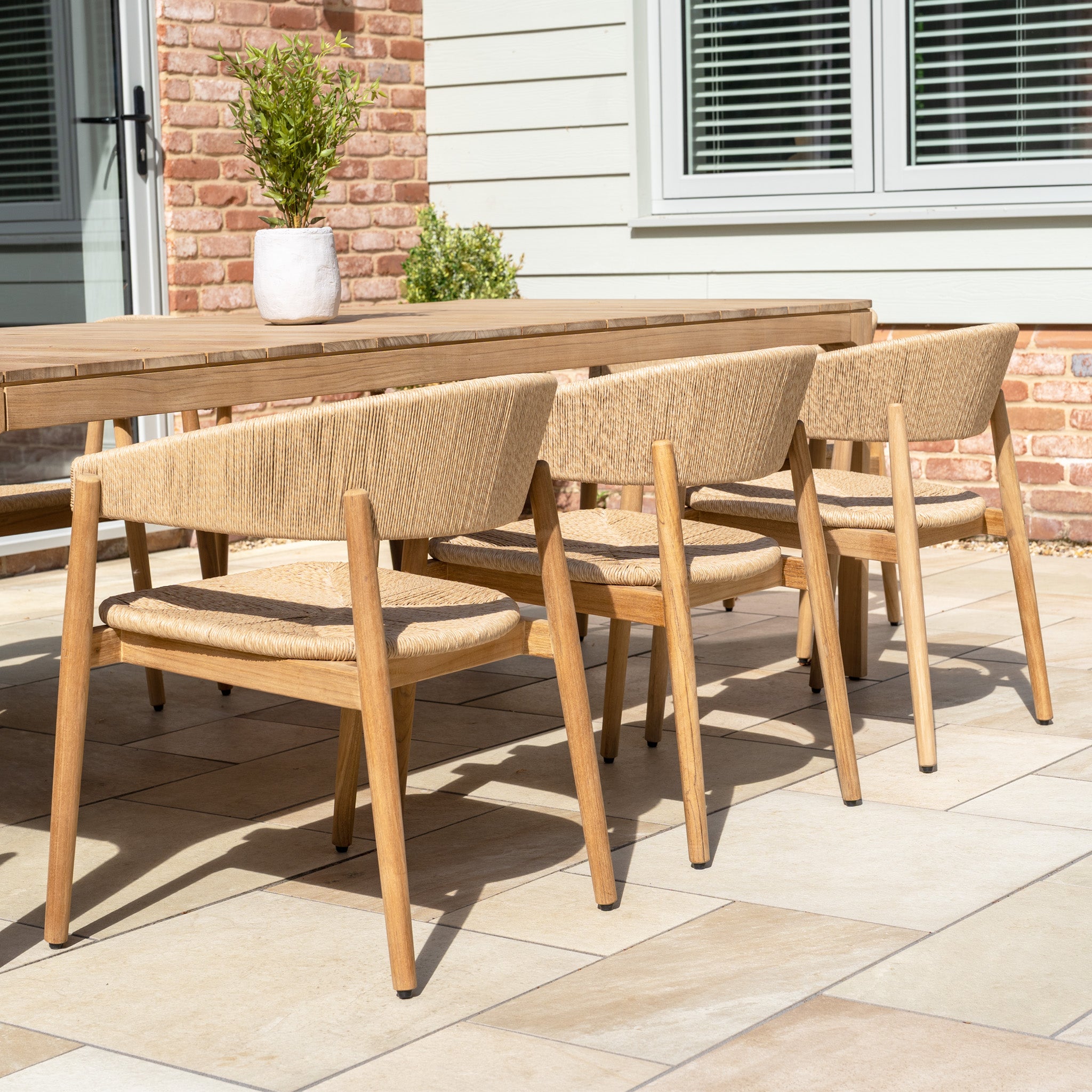Four wooden chairs from the Lucca 8 Seat Dining Set with Teak Table in Natural Weave are arranged around the outdoor dining table on a tiled patio, with a potted plant adding charm to the scene.