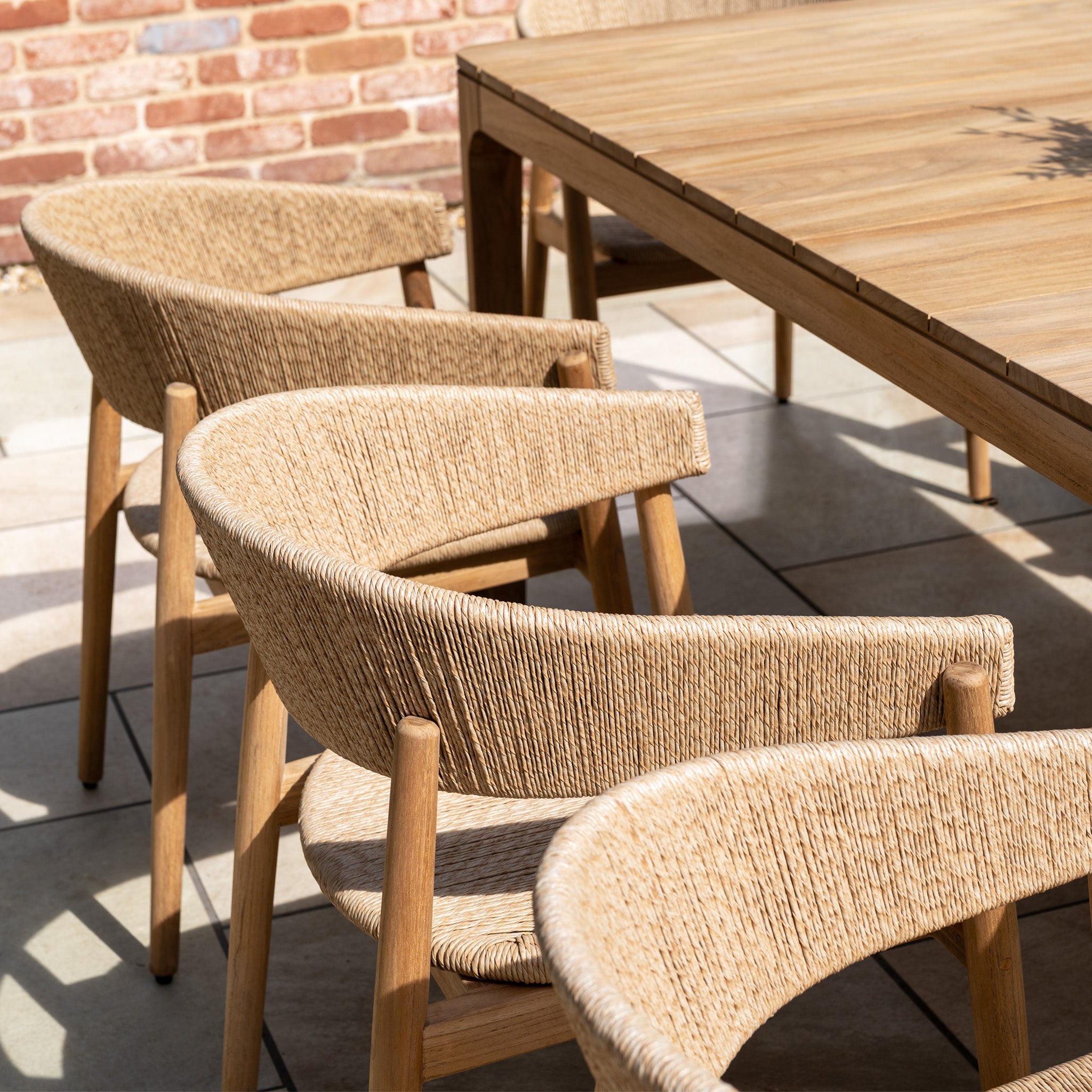 The Lucca 8 Seat Dining Set with Teak Table in Natural Weave sits on a tile patio by a brick wall—stylish wicker chairs and a teak table create an inviting space for elegant outdoor dining.