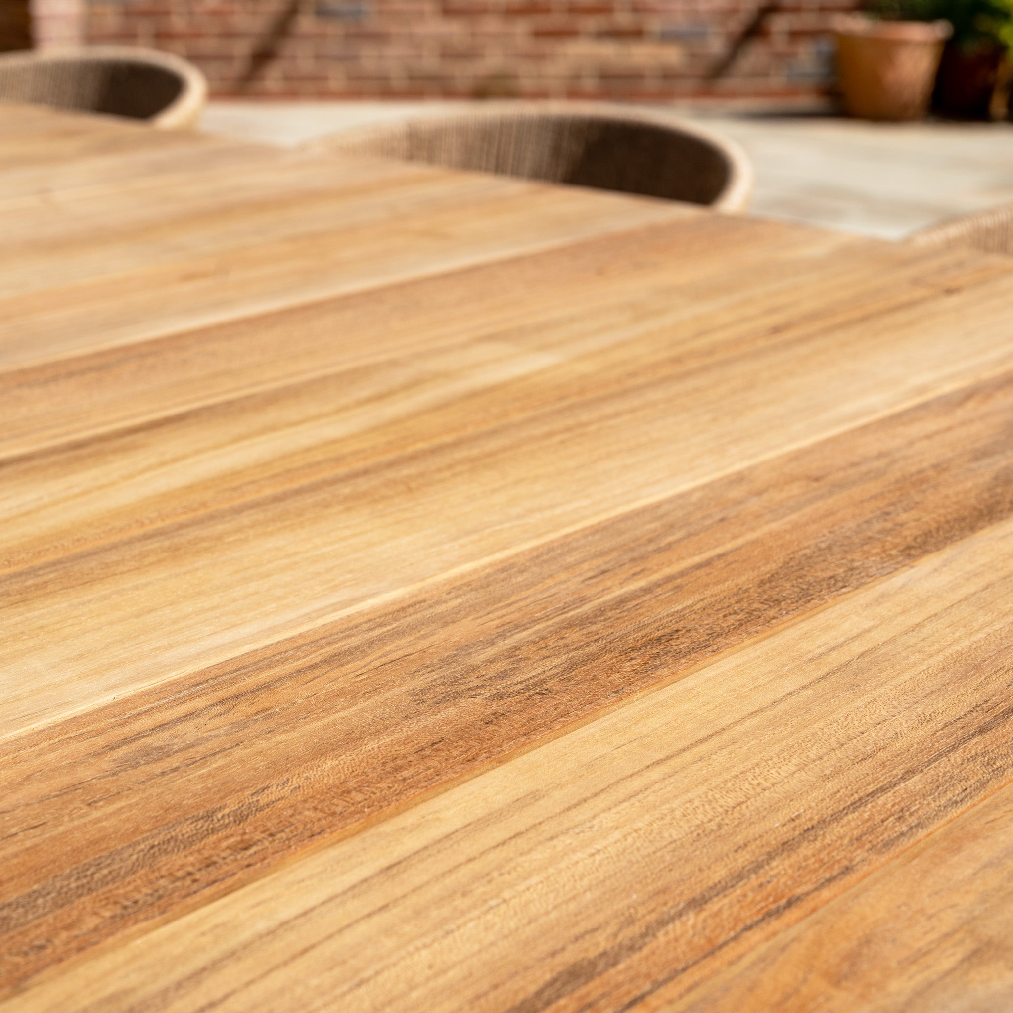 Close-up of the Lucca 8 Seat Dining Set with Teak Table in Natural Weave, featuring hand-woven wicker chairs and softly blurred potted plants in the background.