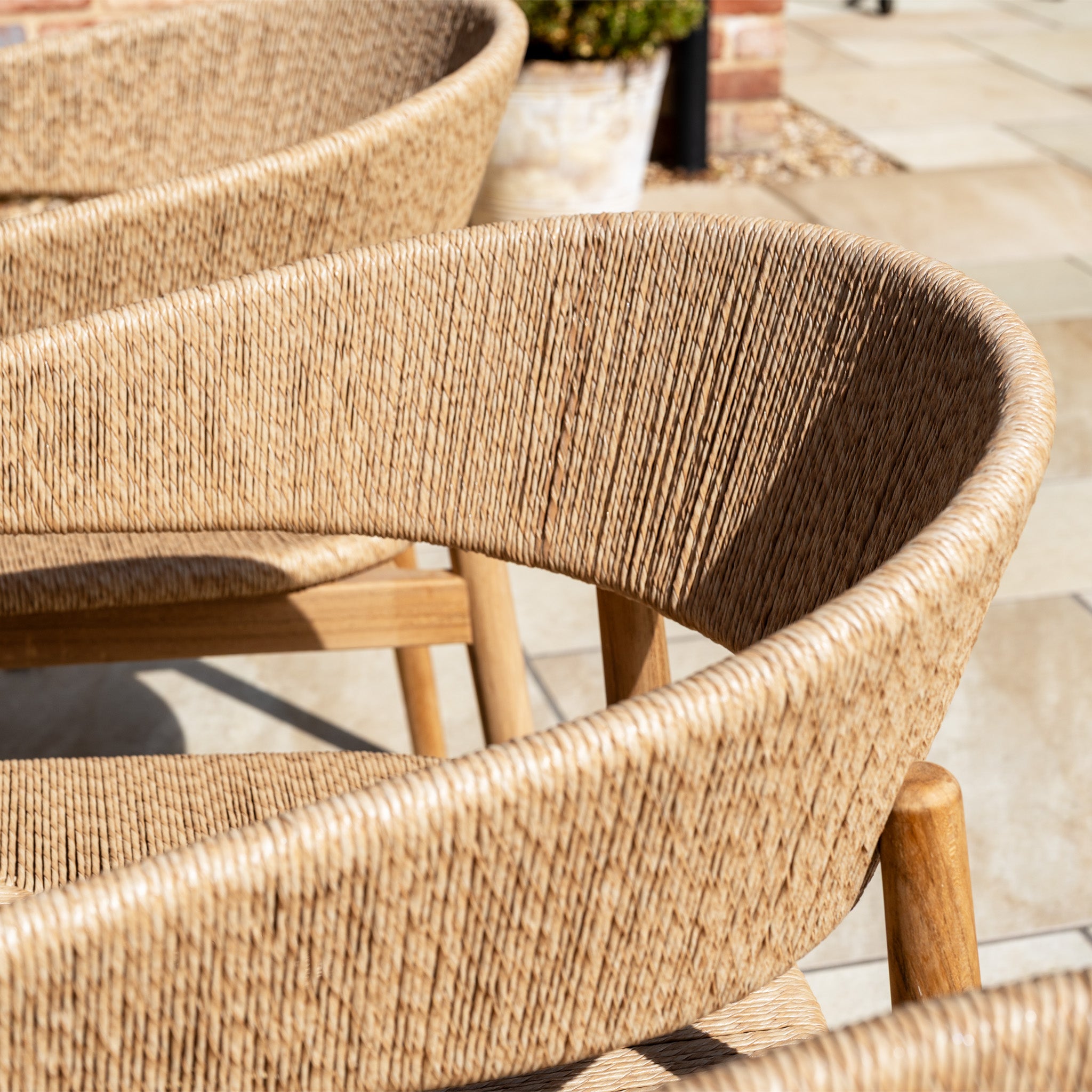 Close-up of woven rattan chairs from the Lucca 8 Seat Dining Set with Teak Table in Natural Weave, featuring curved backs on a sunlit stone patio—ideal for stylish outdoor dining.