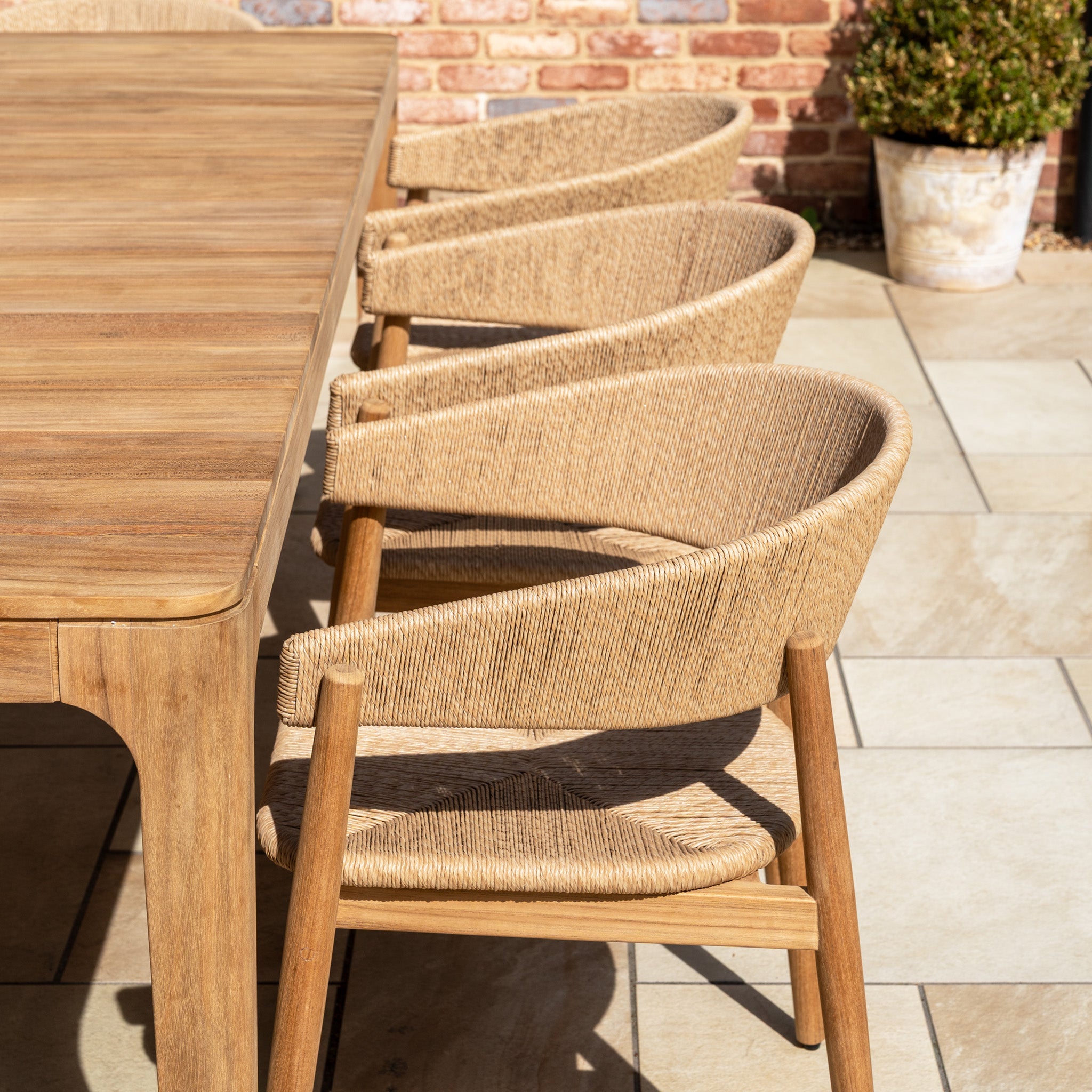 The Lucca 8 Seat Dining Set with Teak Table in Natural Weave features hand-woven wicker chairs around a teak table on a sunlit patio, creating an inviting outdoor dining area with a potted plant and brick wall nearby.
