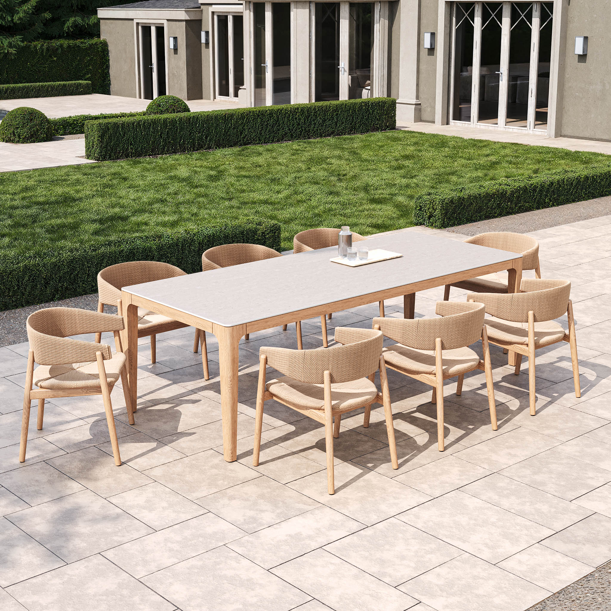 The Lucca 8 Seat Dining Set with Ceramic Top Table in Natural Weave includes a teak frame and ceramic tabletop, accompanied by eight wicker chairs, set on a patio surrounded by trimmed hedges.