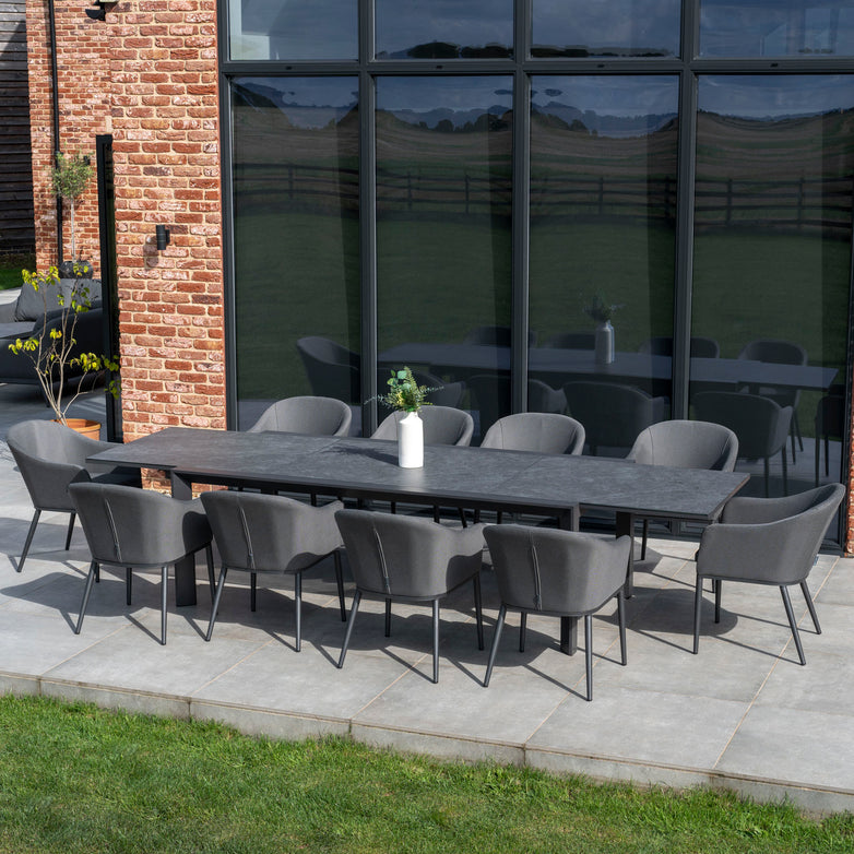 The Luna 10 Seat Outdoor Fabric Extending Ceramic Dining Set in Grey, featuring a long dark table surrounded by eight chairs crafted from durable Luna outdoor fabric, creates a stunning centerpiece on the patio against a brick wall. This stain-proof dining set artfully combines style and functionality for any gathering.