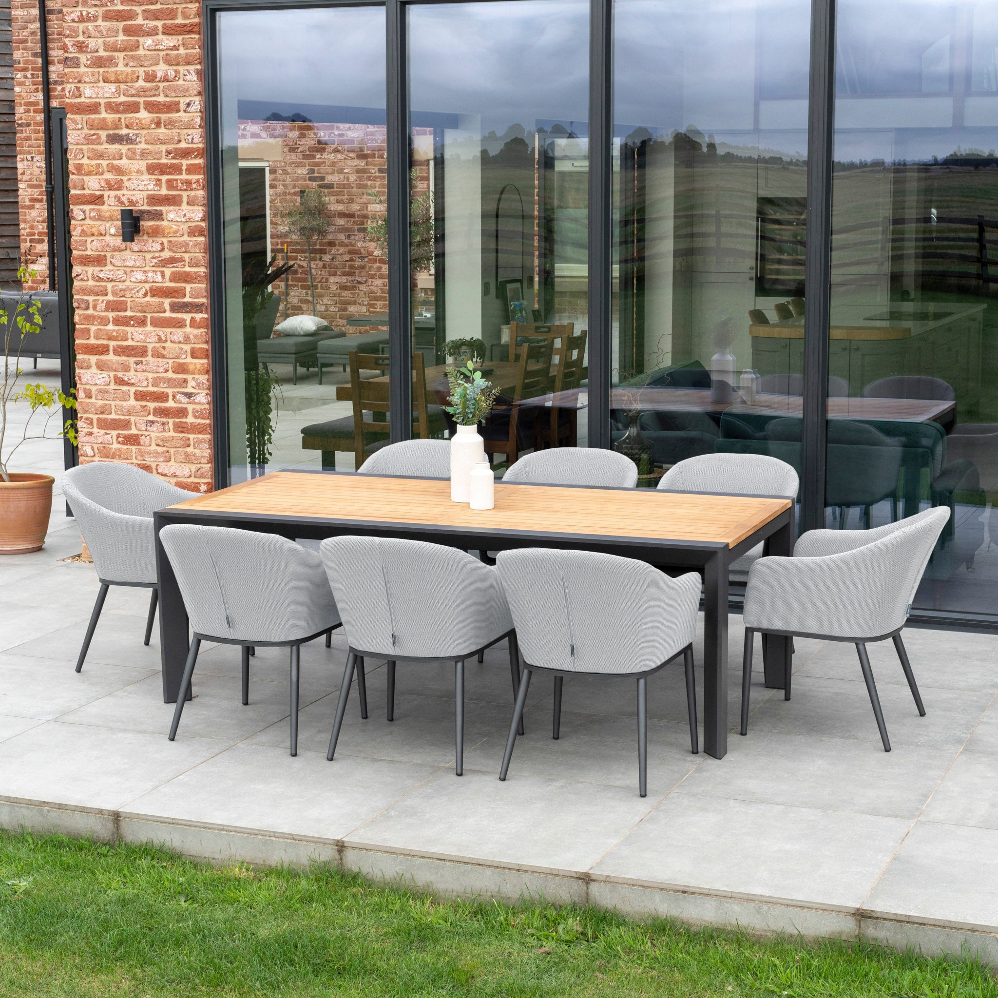 Outdoor patio featuring the Luna 8 Seat Outdoor Fabric Extending Teak Dining Set in Oyster Grey, complete with eight cushioned chairs, set against a modern glass and brick building.