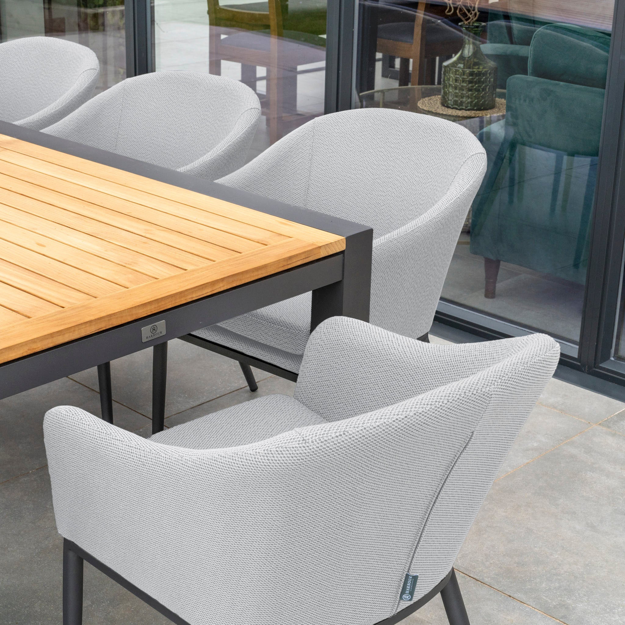 Outdoor dining area featuring the Luna 8 Seat Outdoor Fabric Extending Teak Dining Set in Oyster Grey, with plush grey cushioned chairs positioned near glass doors that reflect additional seating inside, all crafted from maintenance-free materials.
