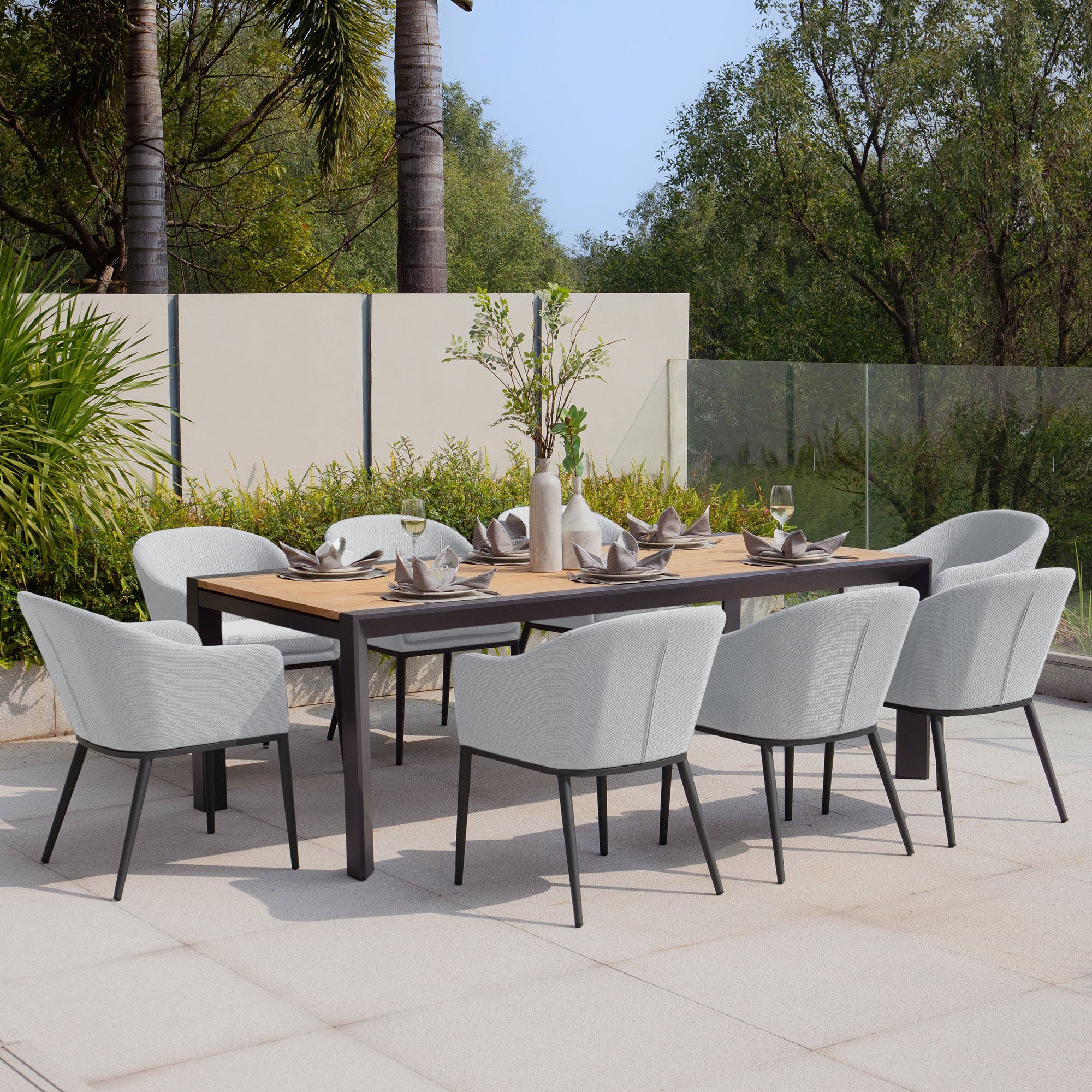 The Luna 8 Seat Outdoor Fabric Extending Teak Dining Set in Oyster Grey features an extendable teak table, eight cushioned chairs made from low-maintenance outdoor fabric, complemented by elegant tableware and a stunning centerpiece, set amidst lush greenery.