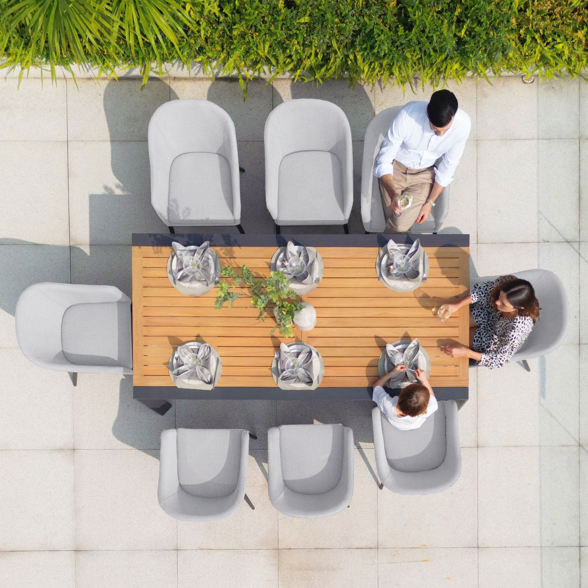 Aerial view of three people sitting at an outdoor dining table set with ten chairs from the Luna 10 Seat Outdoor Fabric Extending Teak Dining Set in Oyster Grey, decorated with plants, and featuring stain-proof Luna outdoor fabric.