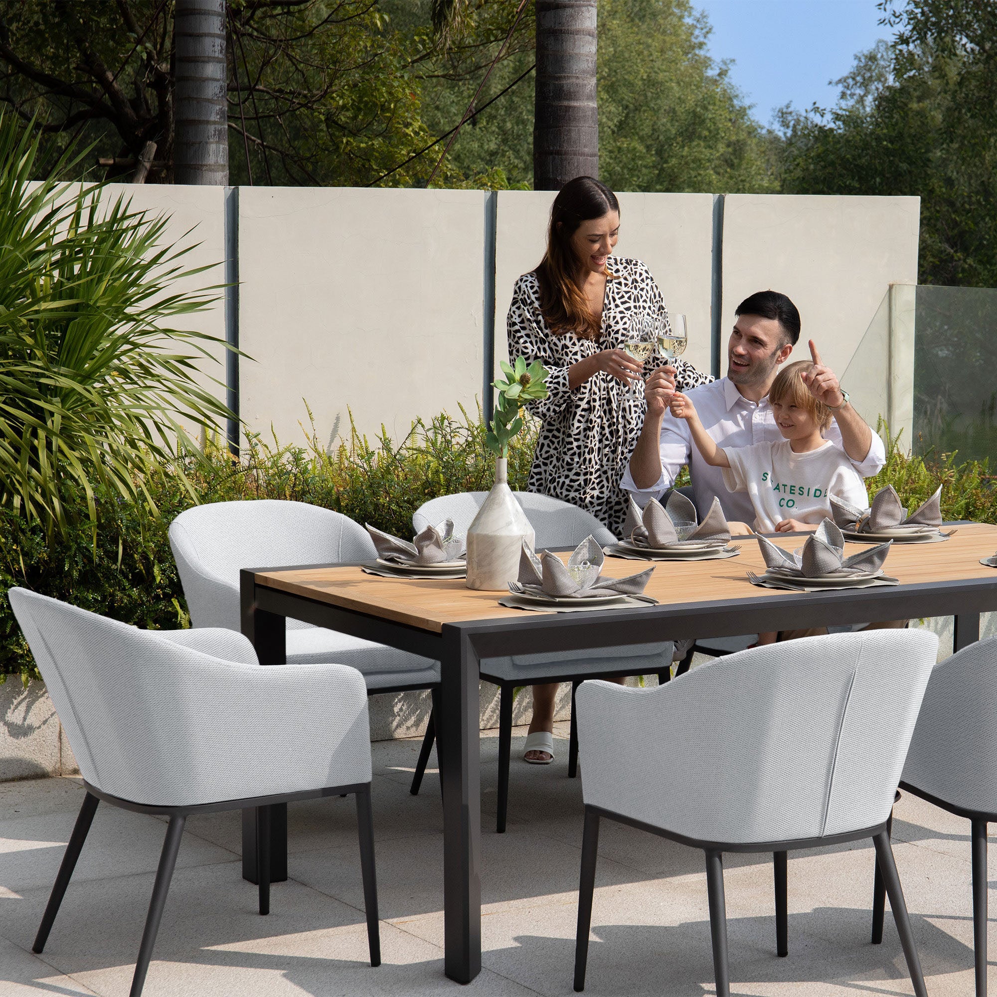 A family of three at an outdoor dining table with greenery and trees in the background, happily interacting around their Luna 8 Seat Outdoor Fabric Extending Teak Dining Set in Oyster Grey.