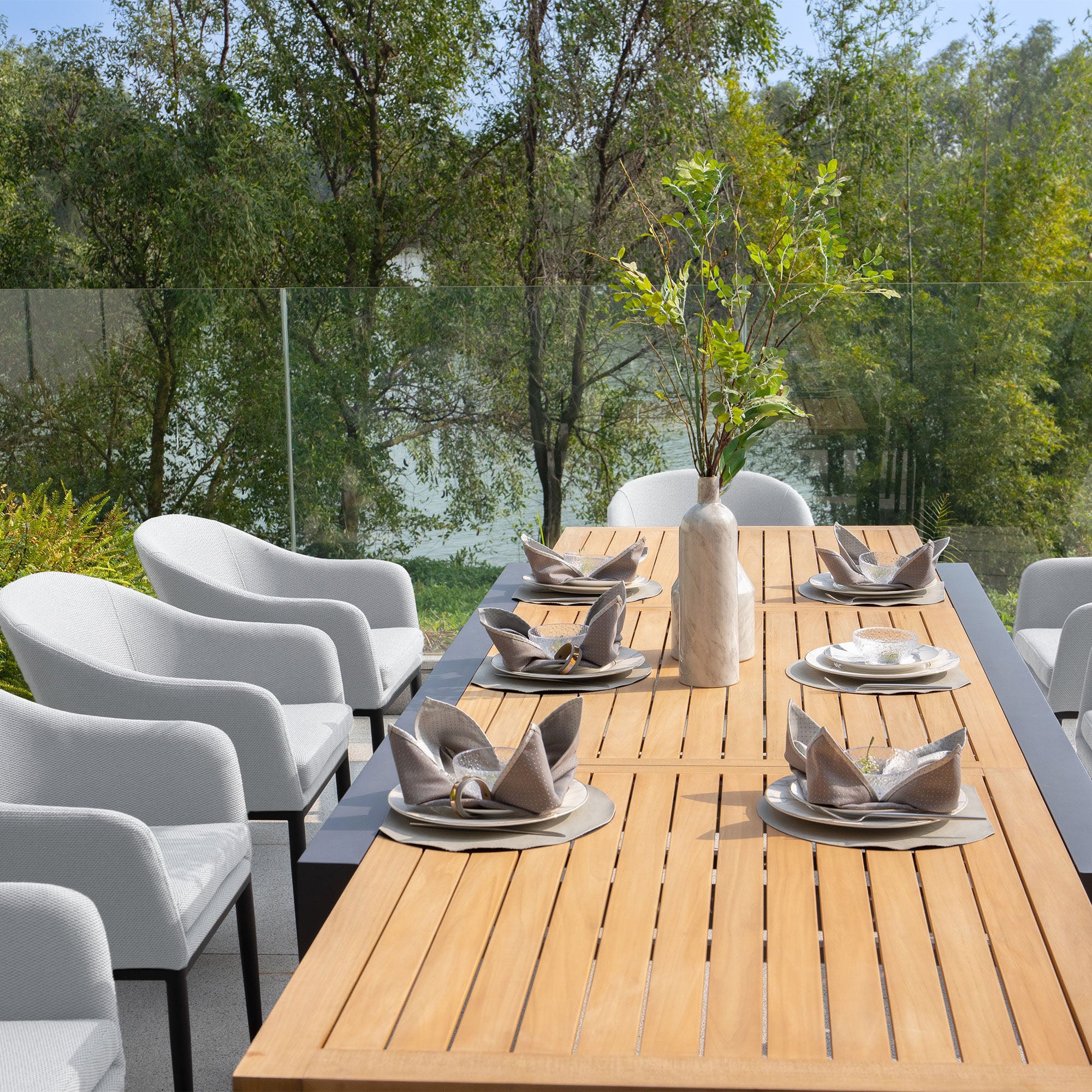 The Luna 8 Seat Outdoor Fabric Extending Teak Dining Set in Oyster Grey, surrounded by lush greenery, is set for six with white chairs, folded napkins, and a vase with branches. This extending teak dining set offers ample space for gatherings while being virtually maintenance-free.