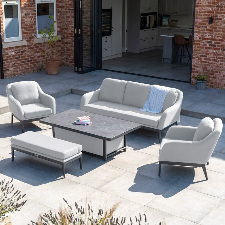 Outdoor patio featuring a Luna 3 Seat Outdoor Fabric Sofa Set with Rising Table in Oyster Grey, two armchairs, a bench, and a central coffee table, with a Harbour Lifestyle brick house in the background.