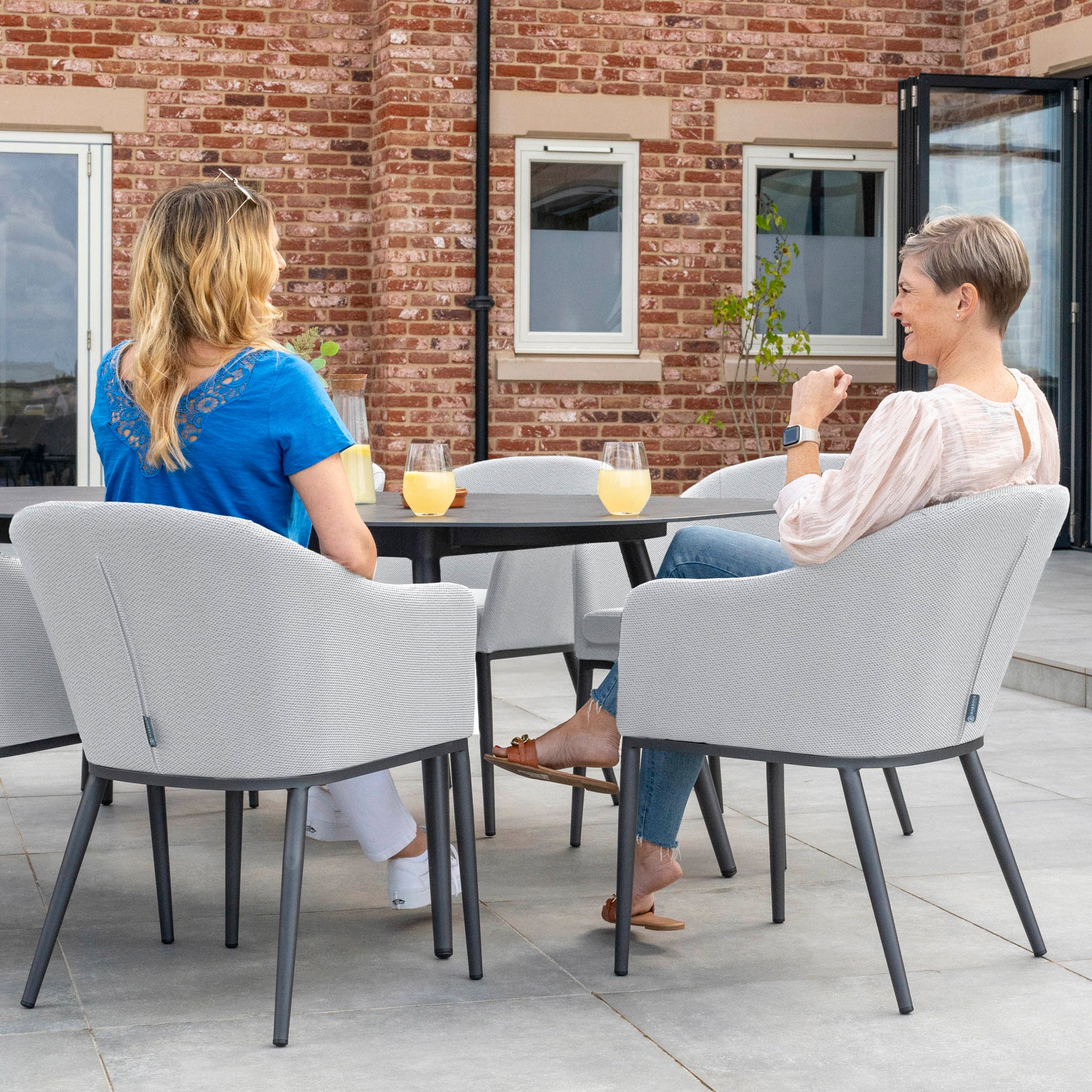 Two women are sitting on the Luna 6 Seat Outdoor Fabric Oval Ceramic Dining Set in Oyster Grey on a patio, enjoying drinks at a table with a brick building in the background. The comfortable Luna outdoor fabric makes their evening even more pleasant.